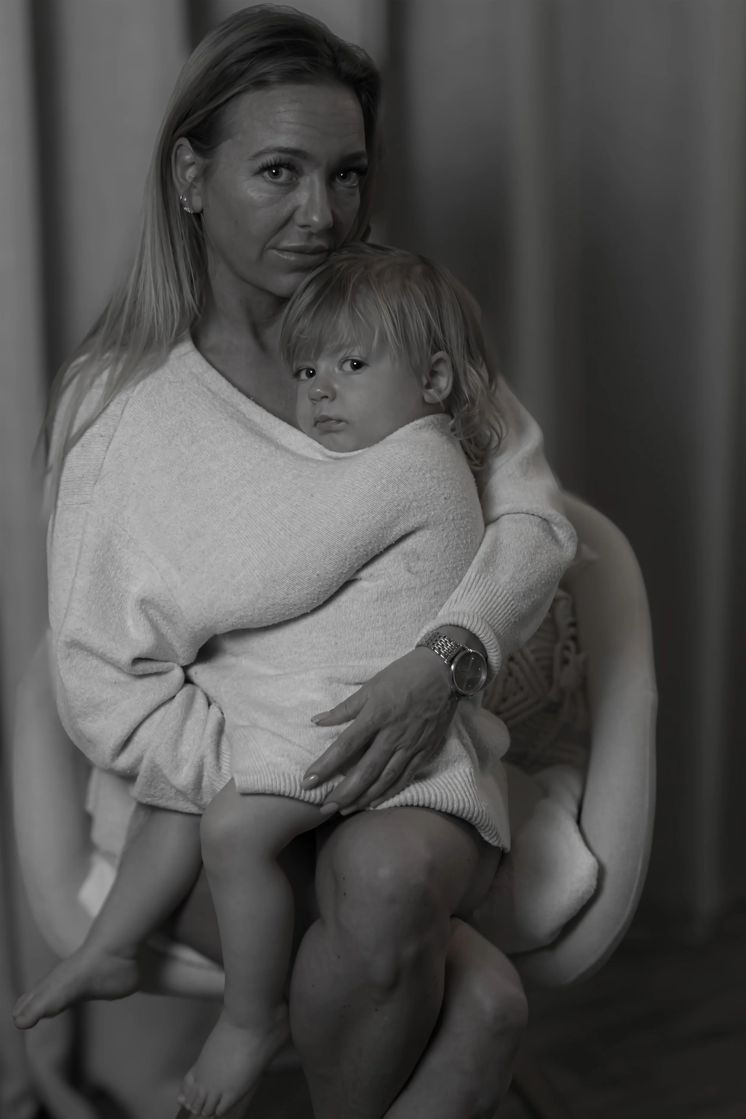 Schwarz-weiß-Fotografie einer alten Frau, die ein kleines Kind auf dem Schoß hält. Die Frau schaut ernst in die Kamera, während das Kind direkt schaut. Sie sitzen auf einem Stuhl, die Umgebung ist unscharf.