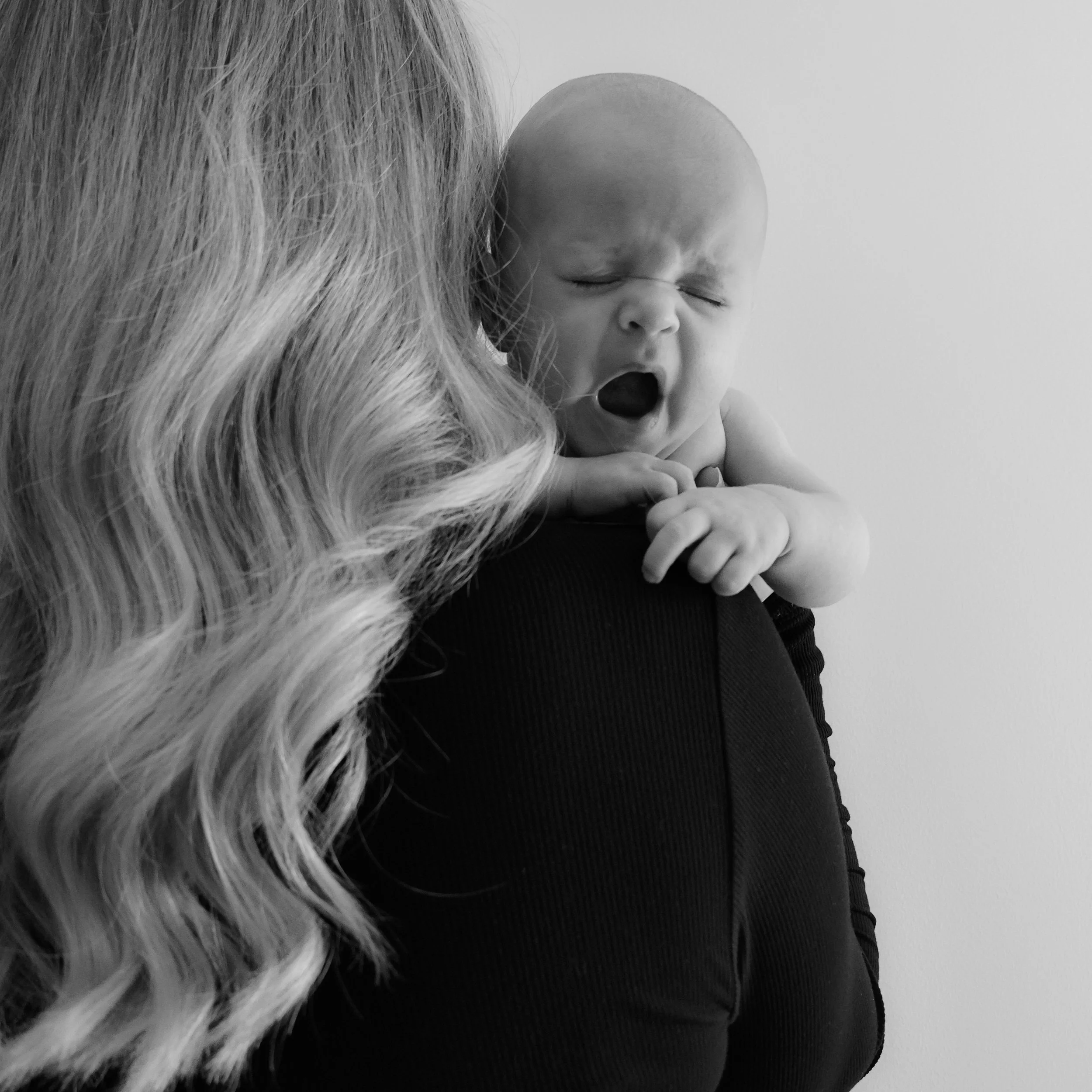 A woman holding a yawning baby close to her shoulder, with the woman's long wavy hair visible and a plain background.