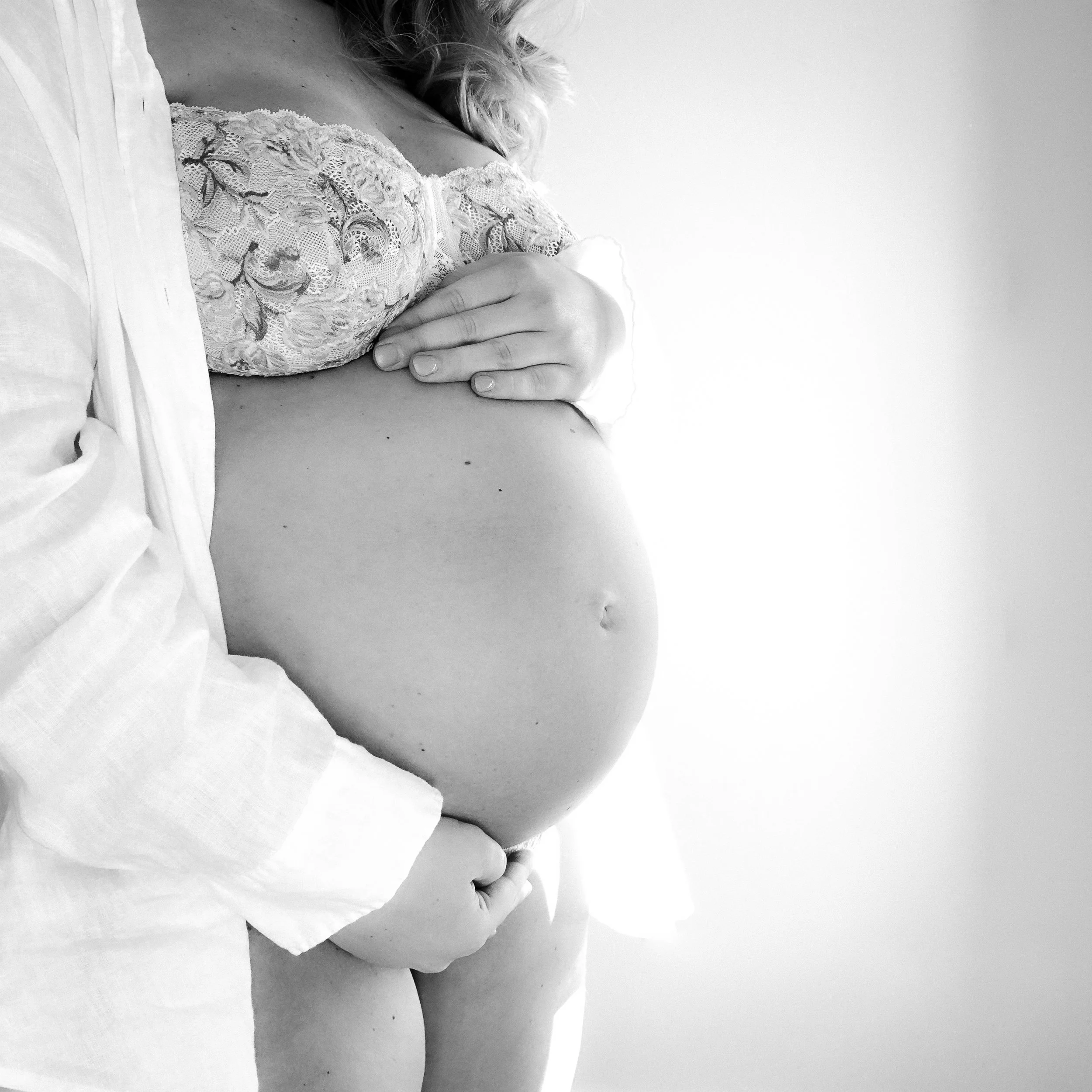 A pregnant woman in a lace top cradling her belly, with a person beside her gently holding her abdomen, in black and white.