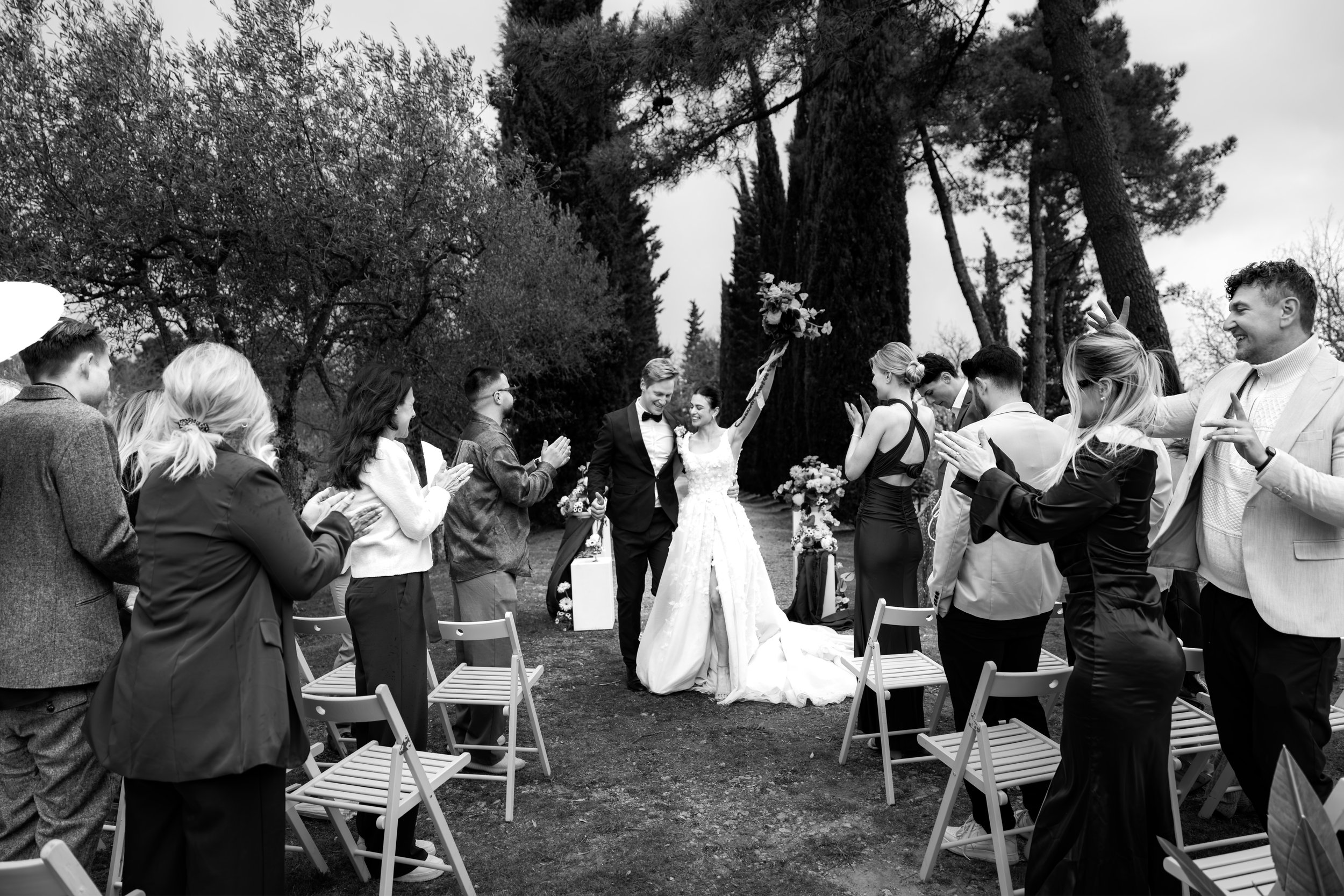 Eine Gruppe von Menschen feiert eine Hochzeit im Freien. Ein Brautpaar steht in der Mitte und alle feiern fröhlich um sie herum.