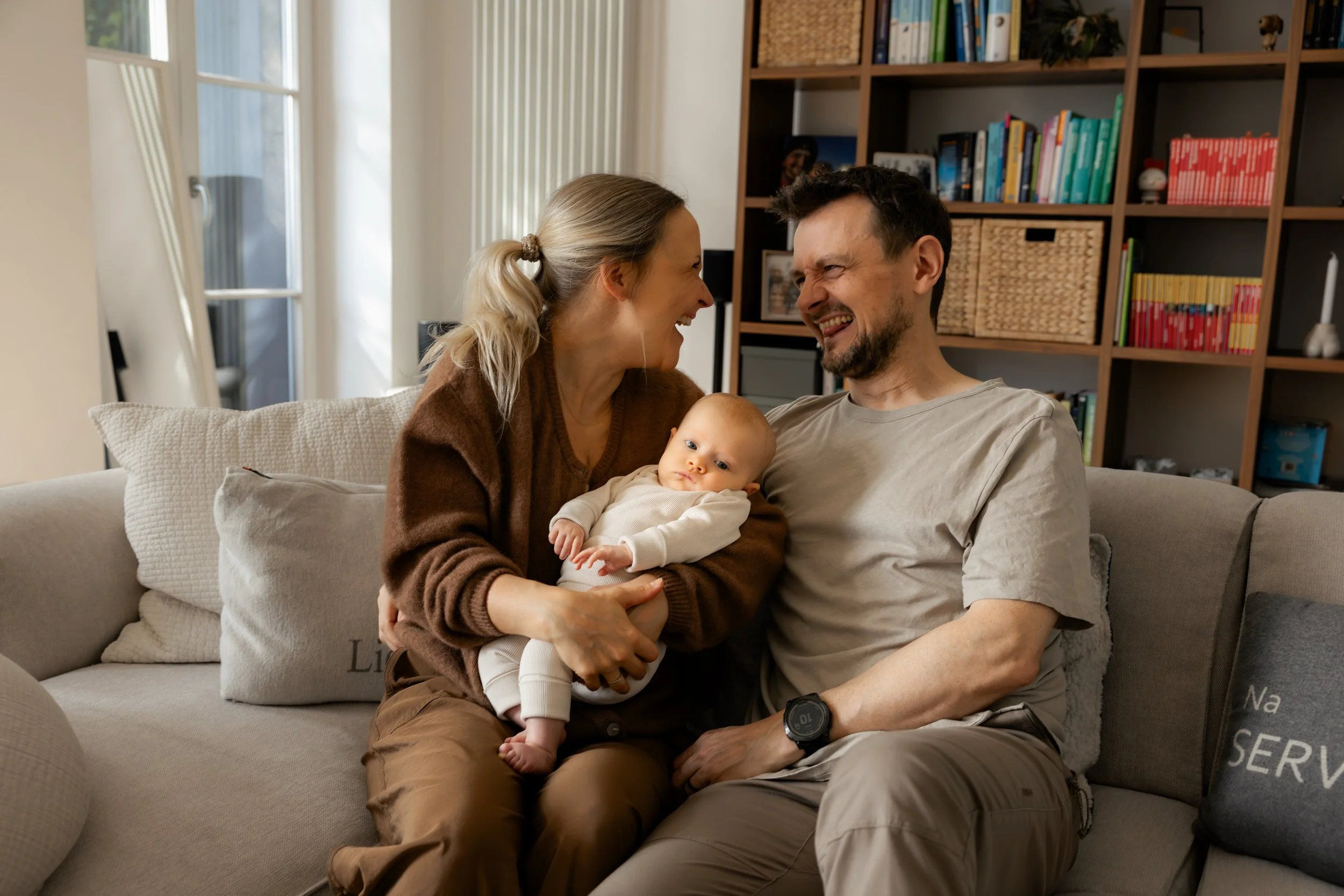 Ein lachendes Paar mit ihrem Baby auf einer Couch im Wohnzimmer, im Hintergrund ein Bücherregal.