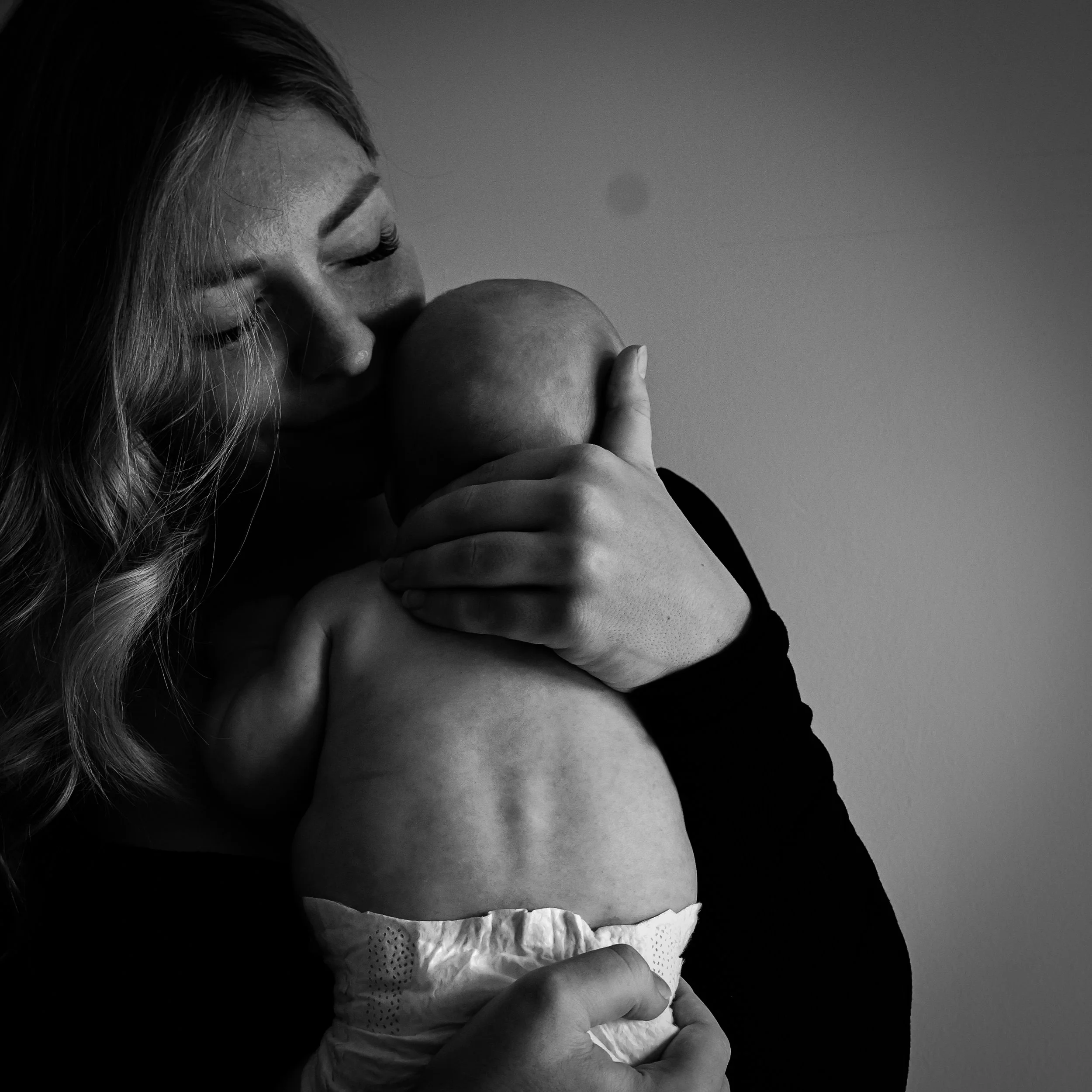 A woman with long wavy hair holding a baby close, both with eyes closed, in a tender moment. The woman is gently kissing the baby's head.