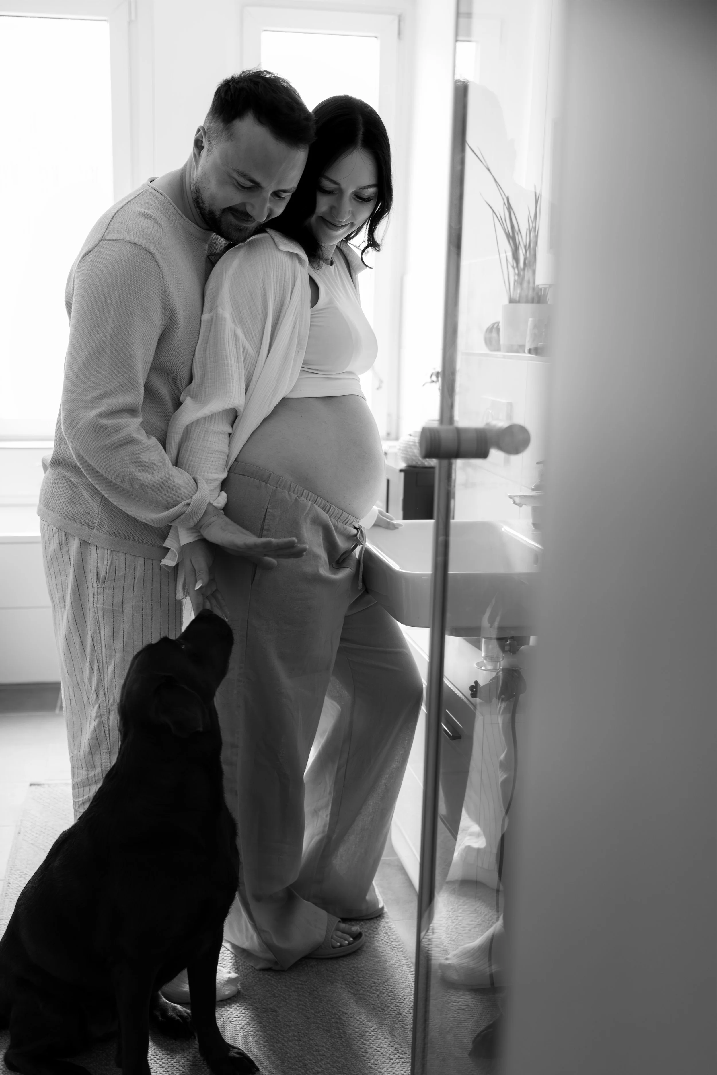 Ein schwangere Frau und ein Mann lachen in einem Badezimmer, während sie auf das Babybauch legen. Ein Hund sitzt auf dem Boden, alle schauen liebevoll auf den Bauch der Frau.