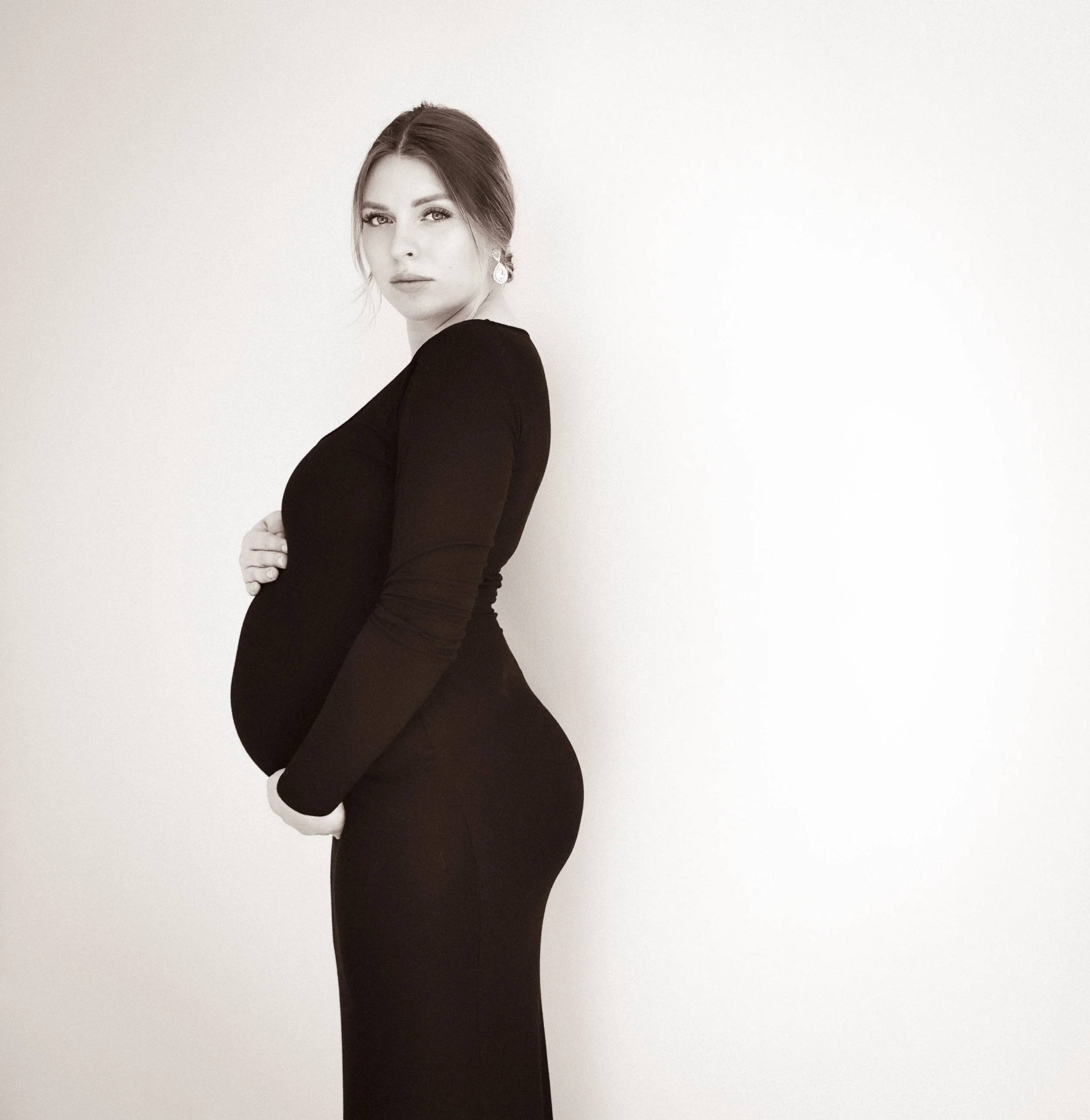 A pregnant woman wearing a black long-sleeve dress, standing sideways with her hands on her belly in front of a plain white wall.