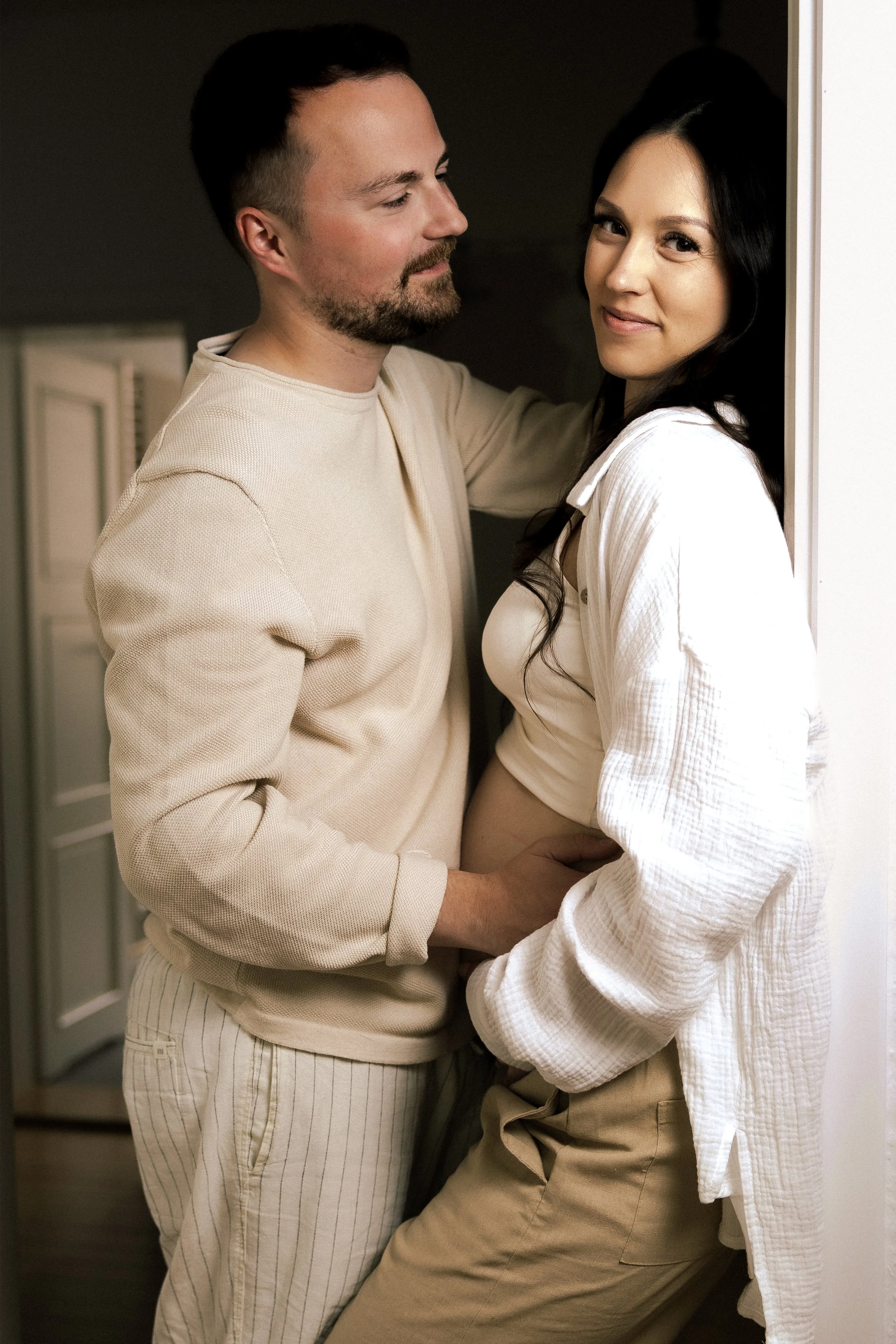 Ein Mann und eine schwangere Frau stehen sich nah in einem Raum, umarmen sich, der Mann lächelt liebevoll, und die Frau schaut direkt in die Kamera.