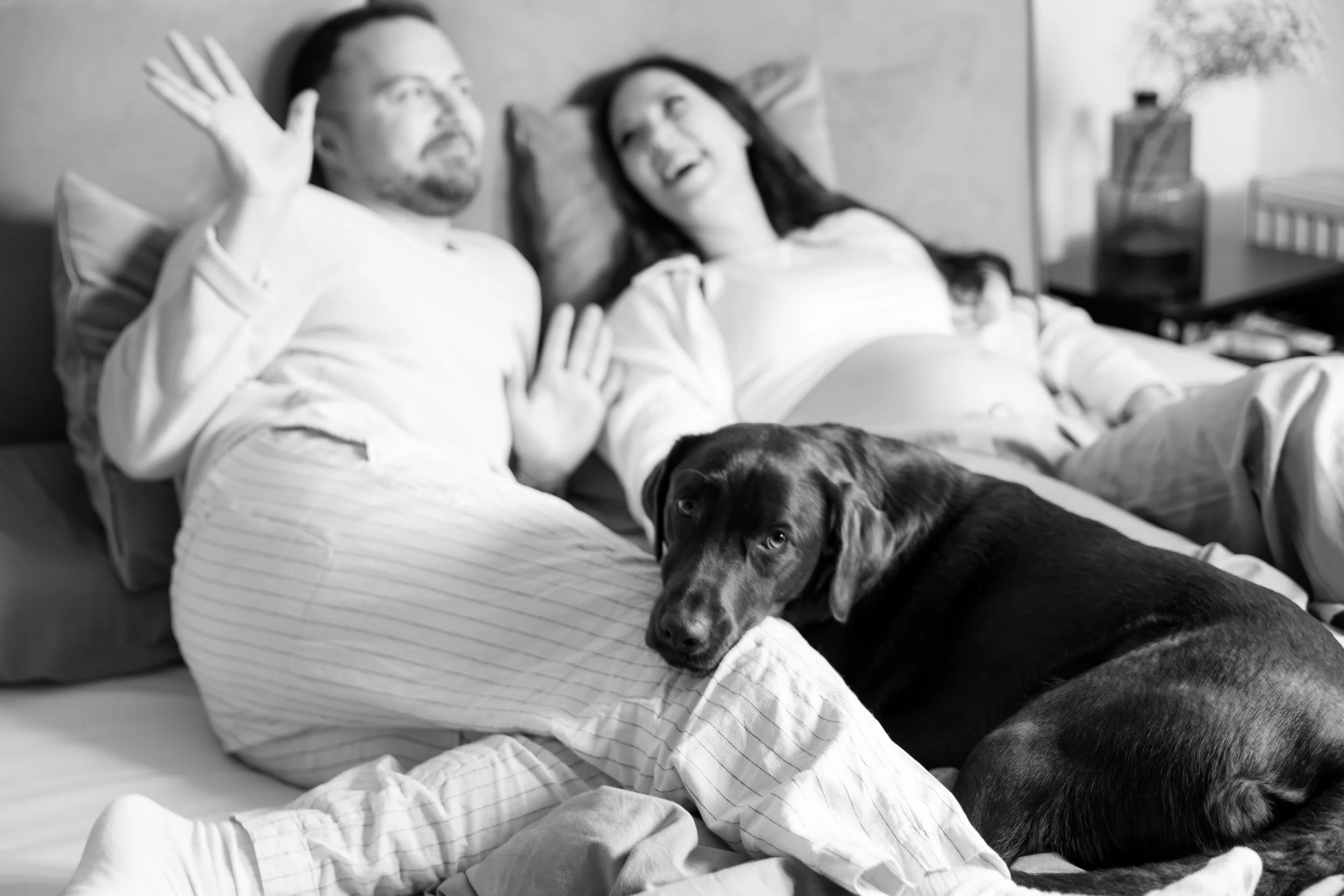 Zwei Menschen liegen auf einem Bett, der Mann winkt, die Frau lacht. Ein Hund liegt auf dem Bett und schaut in die Kamera.