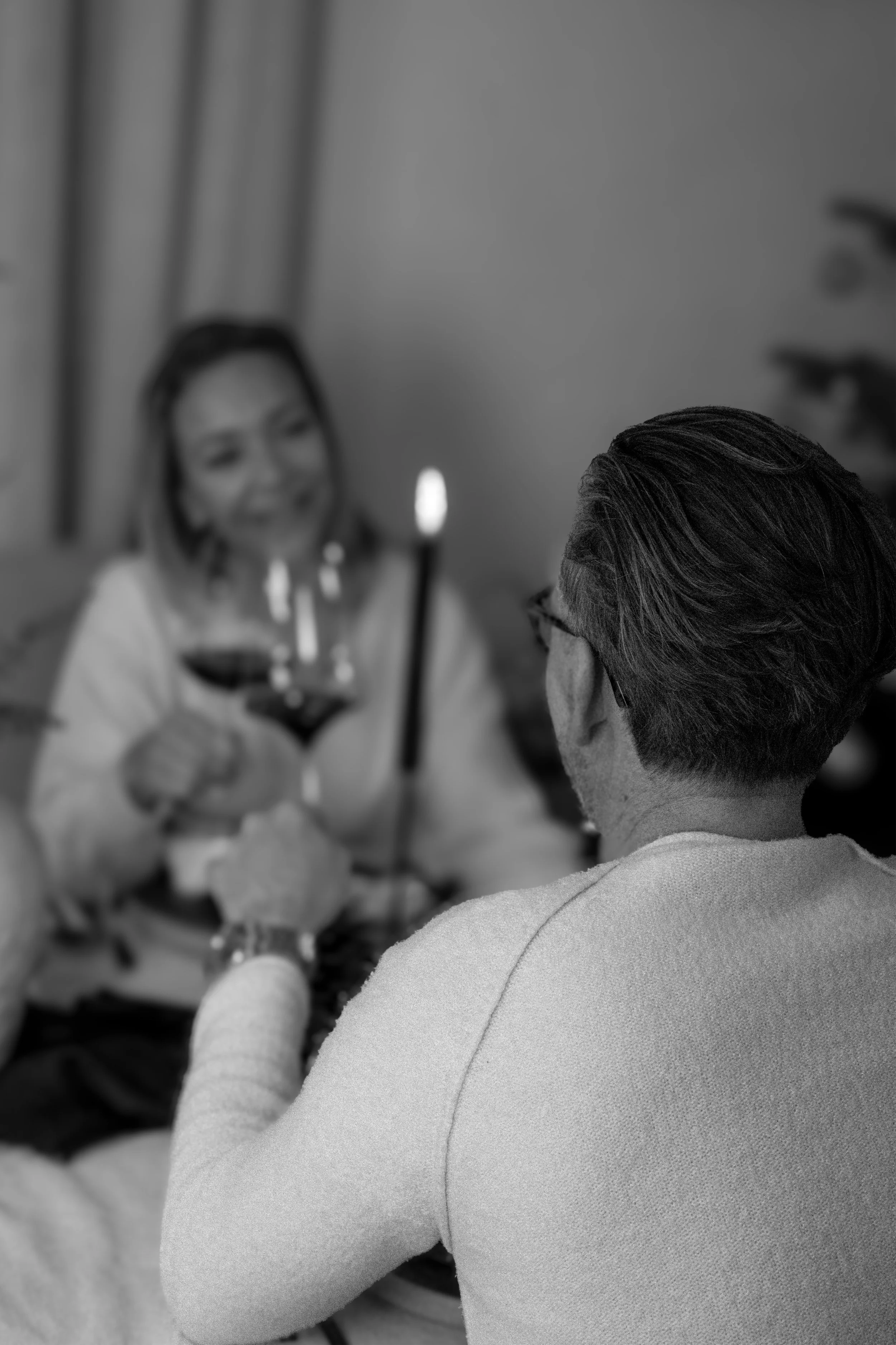 Zwei Personen sitzen sich in einem Raum gegenüber. Die Frau links lächelt und hält ein Glas Wein in der Hand, während der Mann rechts nur von hinten sichtbar ist. Es ist eine schwarze und weiße Aufnahme.
