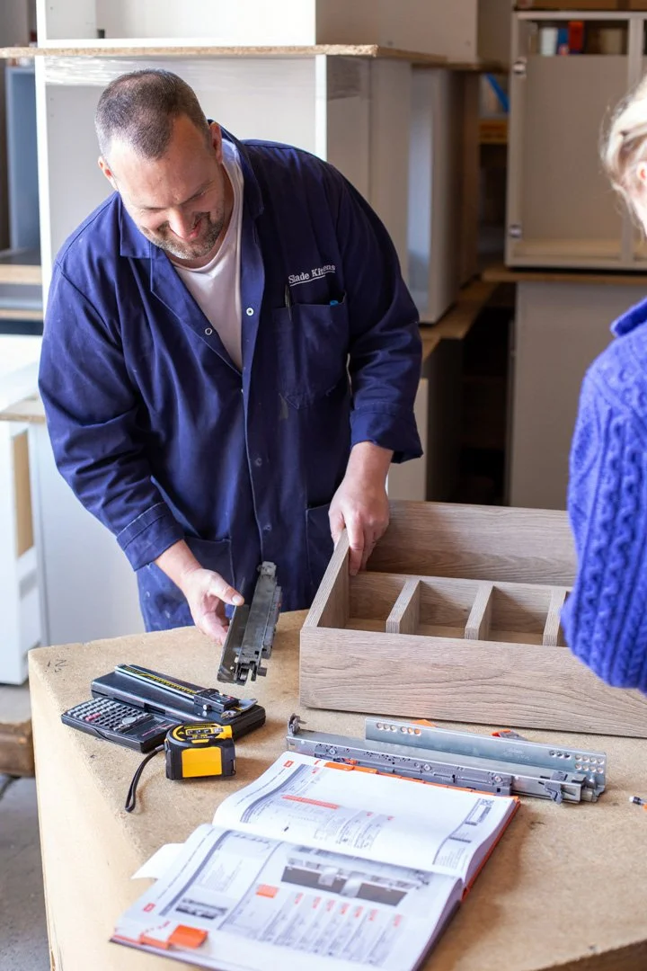 A man works on assembling a wooden drawer with a young girl observing, in a woodworking workshop.