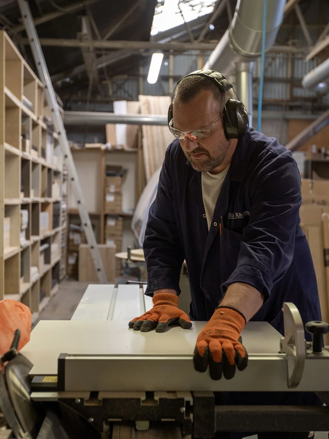 A woodworking shop with a man using a table saw, wearing safety glasses a navy blue work shirt, orange gloves, and hearing protection. Shelves and boxes with wood materials in the background.