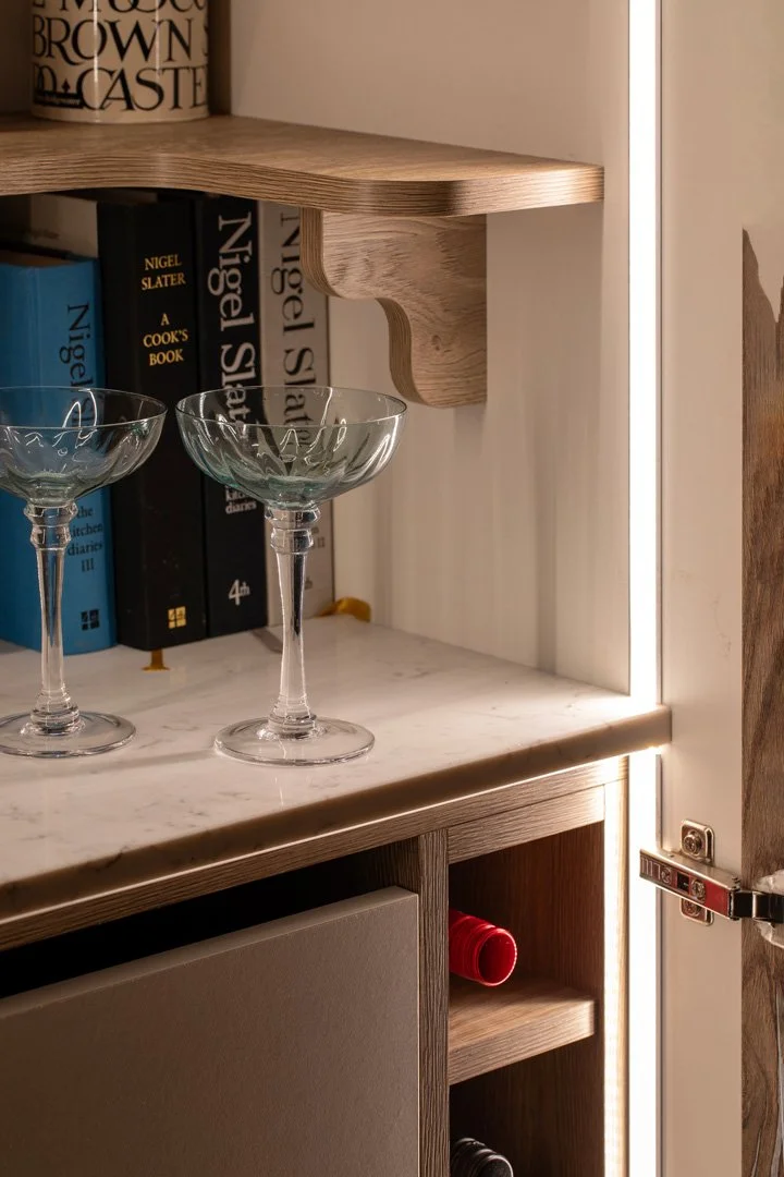 Close-up of a kitchen or bar cabinet with glassware, books, and a shelf with a decorative wooden bracket, illuminated by LED strip lighting.