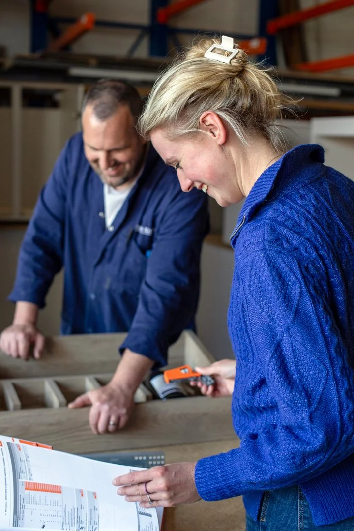 A woman in a blue sweater is laughing and holding a tape measure while looking at a wooden storage unit, with a man in a blue work uniform smiling and looking down in the background.