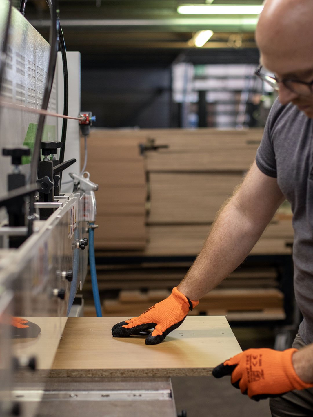 A person wearing orange gloves and glasses operating woodworking machinery in a workshop, with stacks of wood in the background.