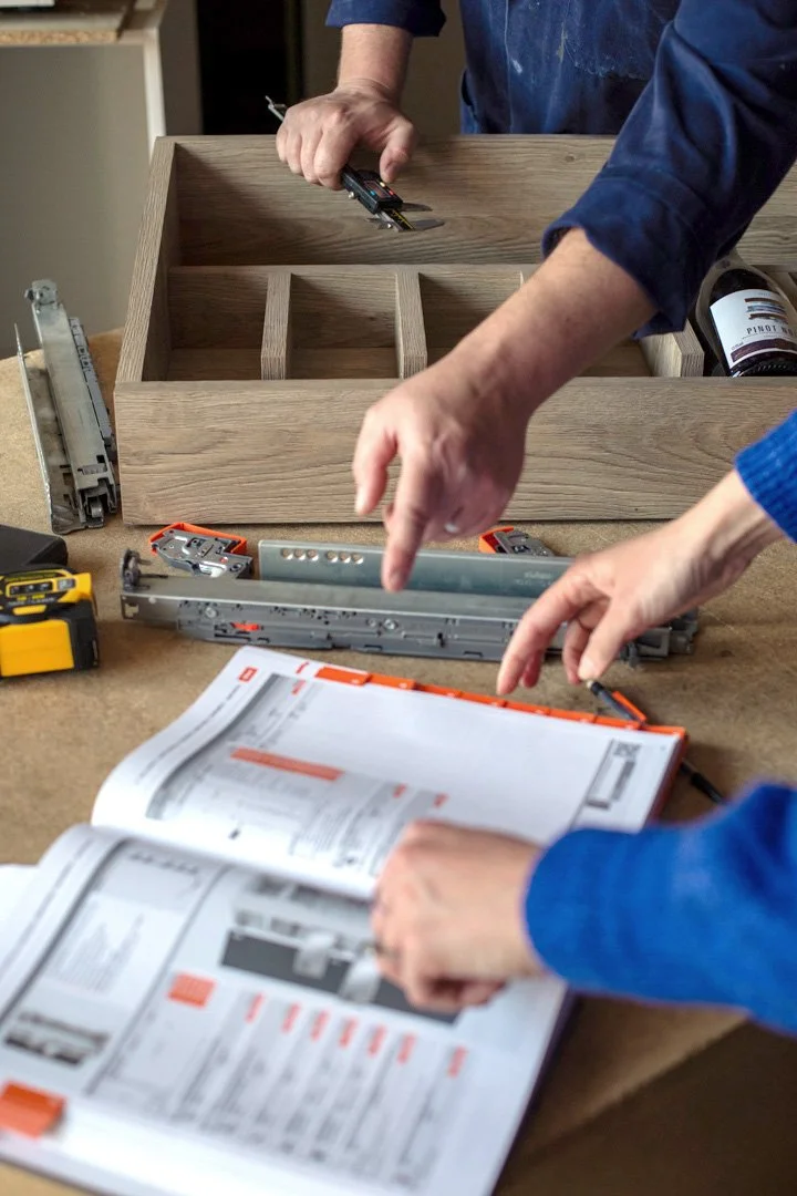 Two people assembling a wooden drawer, with tools, instructions, and hardware on a workbench.