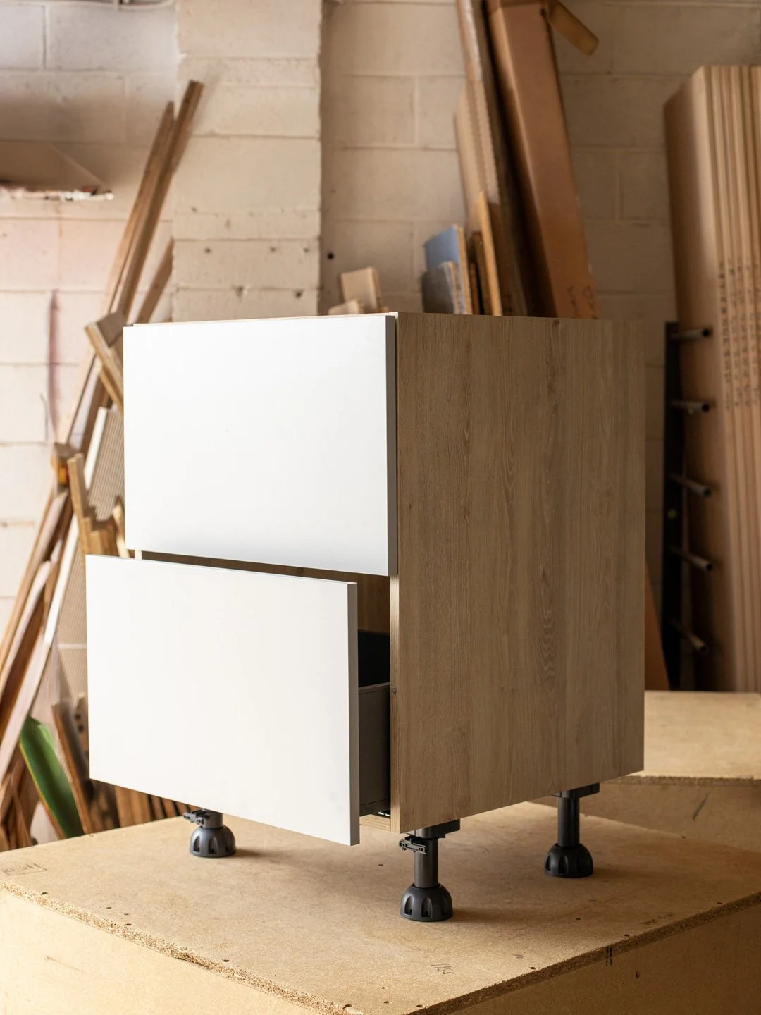 A modern wooden cabinet with two white drawers, supported by adjustable black legs, placed on a wooden surface in a workshop setting.