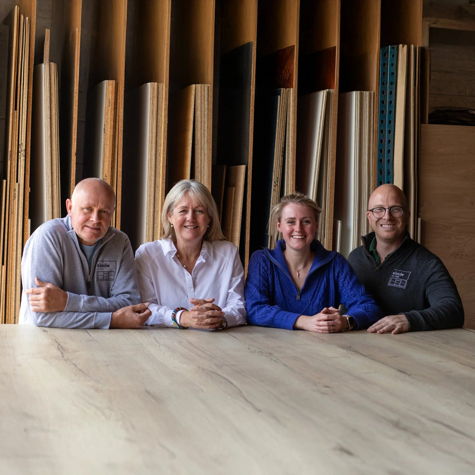 Four people sitting at a wooden table in a woodworking shop, with wooden planks and materials in the background.