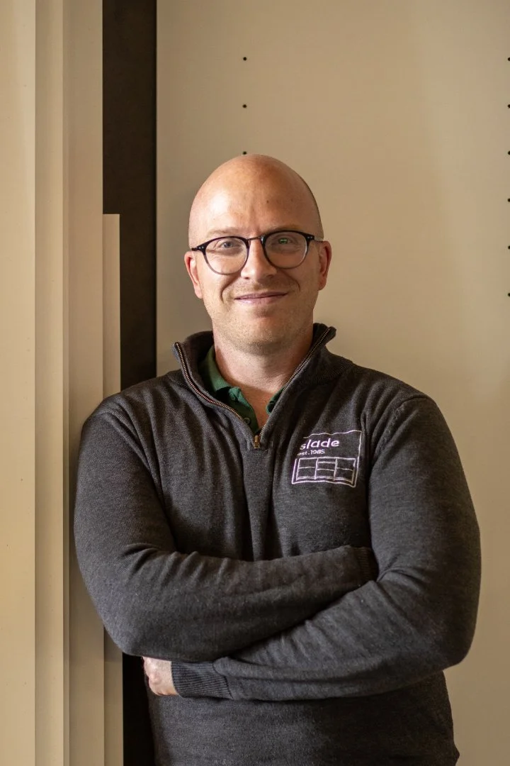 A bald man wearing glasses and a dark gray zip-up sweater with a logo, standing with arms crossed against a light-colored wall.