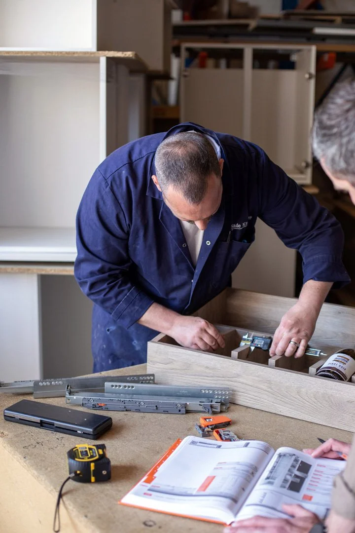 A man in a dark blue shirt assembling furniture at a workshop table, with tools and an open instruction manual nearby.