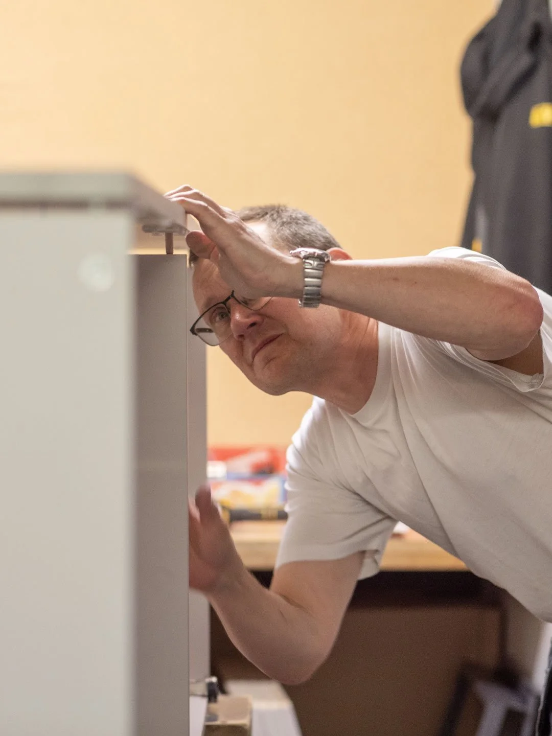 Man inspecting or working inside a cabinet or shelf, wearing glasses and a white t-shirt.