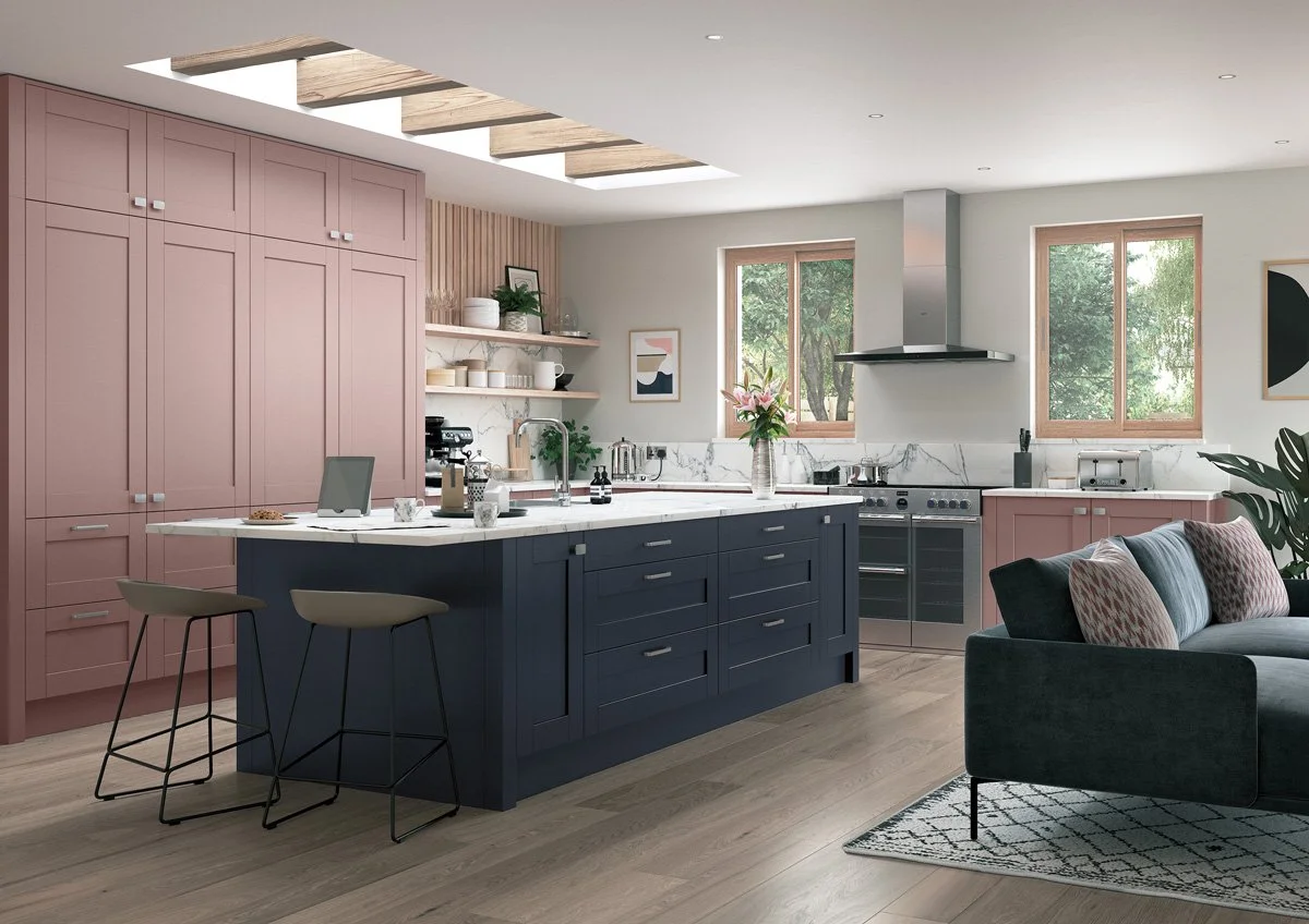 Modern kitchen with pink and navy cabinetry, marble countertops, two windows, open shelving, a centered island with bar stools, and a gray sofa with patterned cushions in the living area.