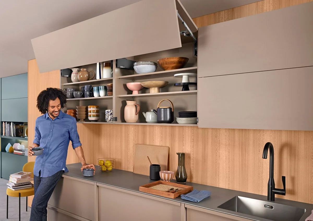 A person in a blue shirt and grey pants in a modern kitchen, smiling, holding a bowl and standing near open shelves filled with various bowls, cups, and decorative items.