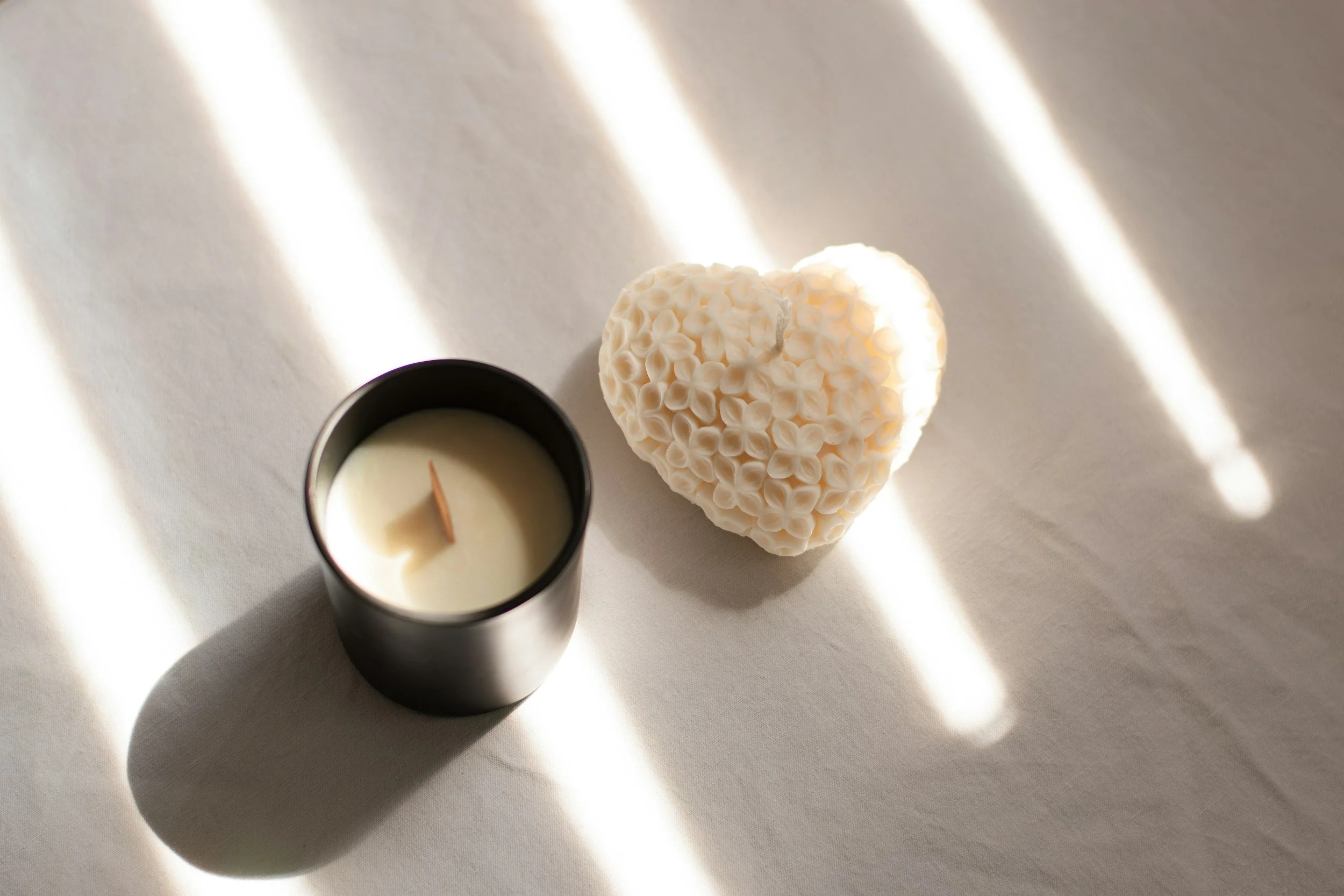 A black candle holder with a white candle, a cream-colored decorative heart with floral pattern, on a white surface with sunlight and shadows.