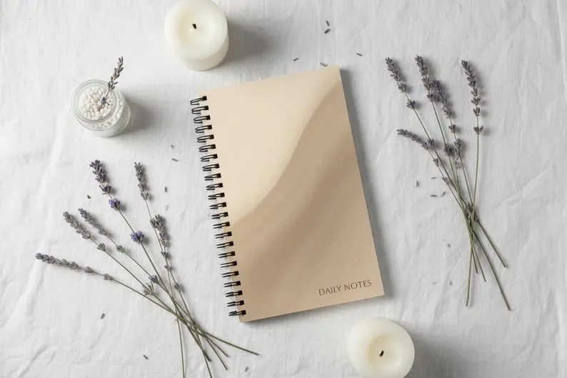 A beige daily notes notebook surrounded by lavender sprigs, two white candles, and a small jar with a lavender sprig inside on a white fabric background.