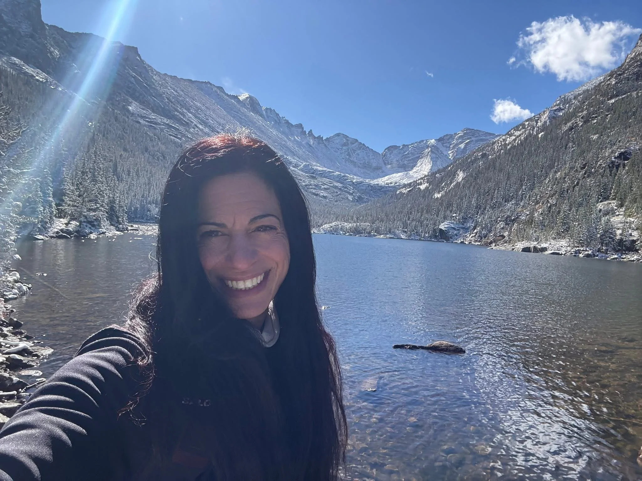 A woman taking a selfie by a mountain lake with snow-covered peaks, evergreen trees, and a clear blue sky in the background.