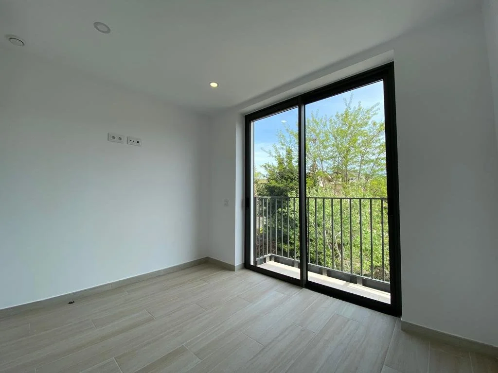 Habitación vacía con piso de cerámica, paredes blancas y una gran ventana de vidrio con balcón, que muestra árboles y cielo azul afuera.