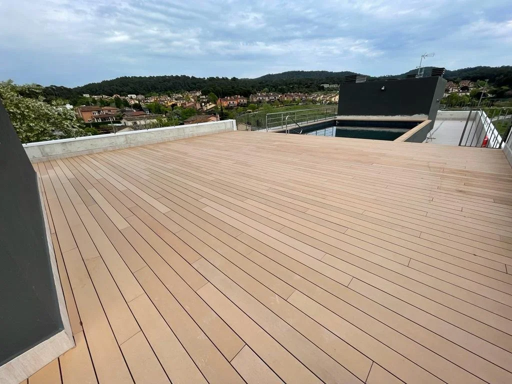 Terraza con piso de madera y vista a áreas verdes y casas en un fondo, con un mural negro y barandillas de metal.