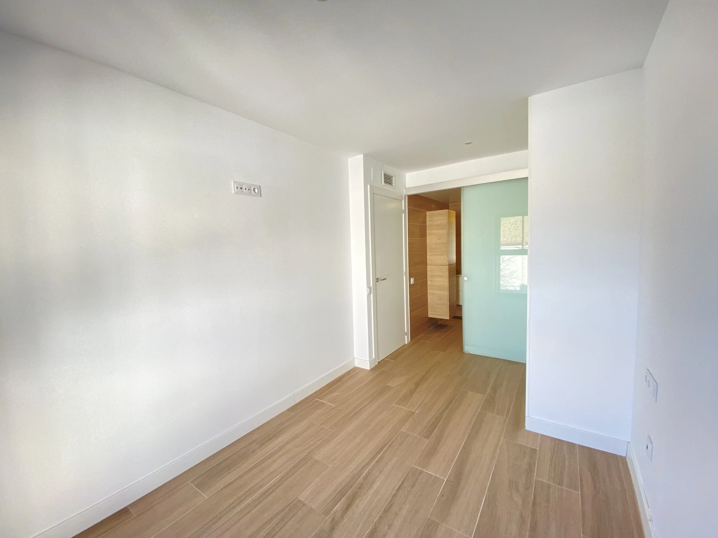 Sala vacía con paredes blancas, piso de madera clara, una puerta de madera y una pared de cristal que deja pasar luz natural.