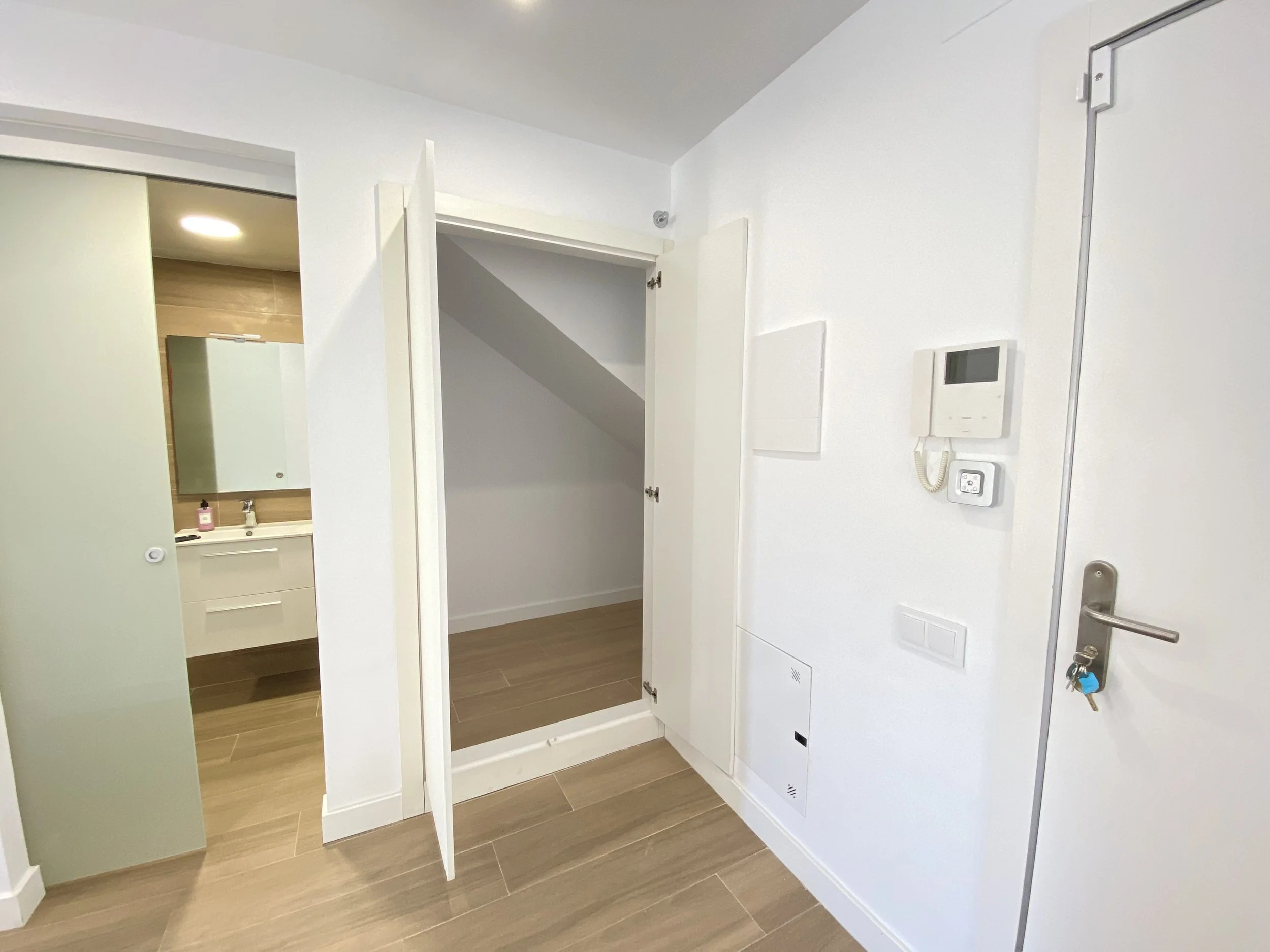 Vista de una entrada de apartamento con pared blanca, una caja de telecomunicaciones, un intercomunicador y una cerradura en la puerta de entrada, con piso de madera y un armario en un rincón.