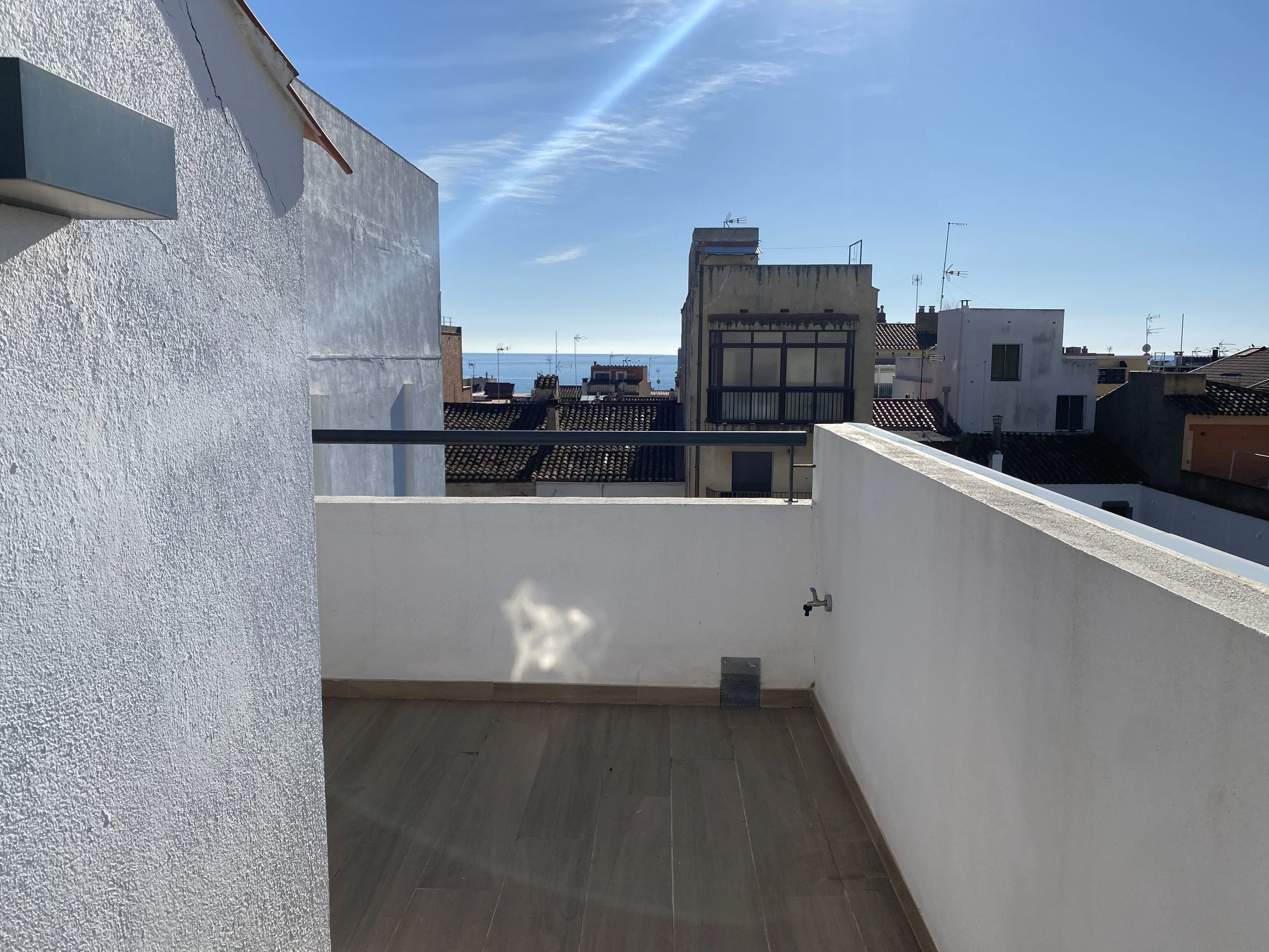 Vista desde una terraza en la ciudad, con varias construcciones de edificios, tejas de techos y el mar en el fondo bajo un cielo despejado.