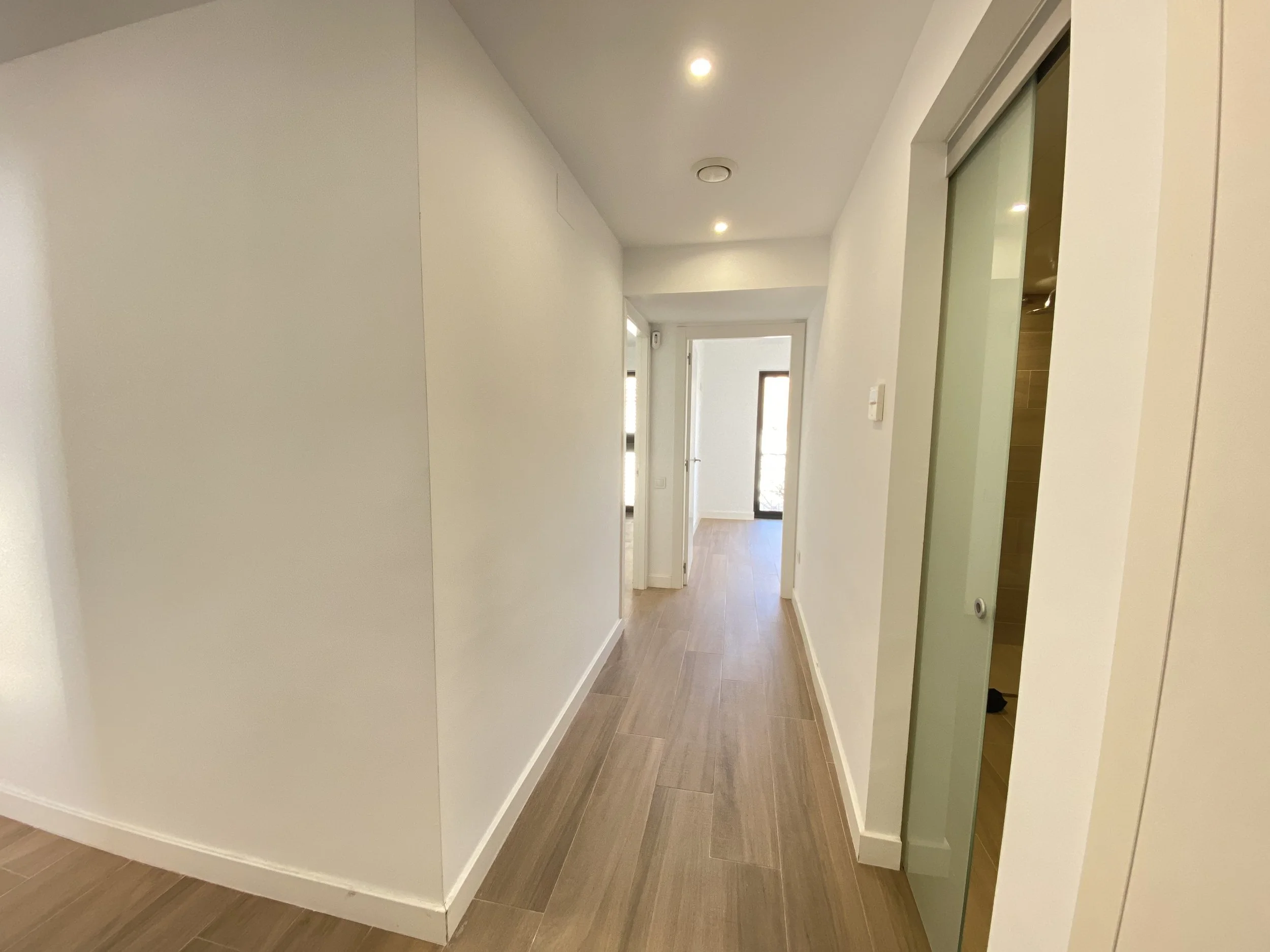 Pasillo interior de una casa moderna con paredes blancas, suelo de madera y puertas en el fondo.