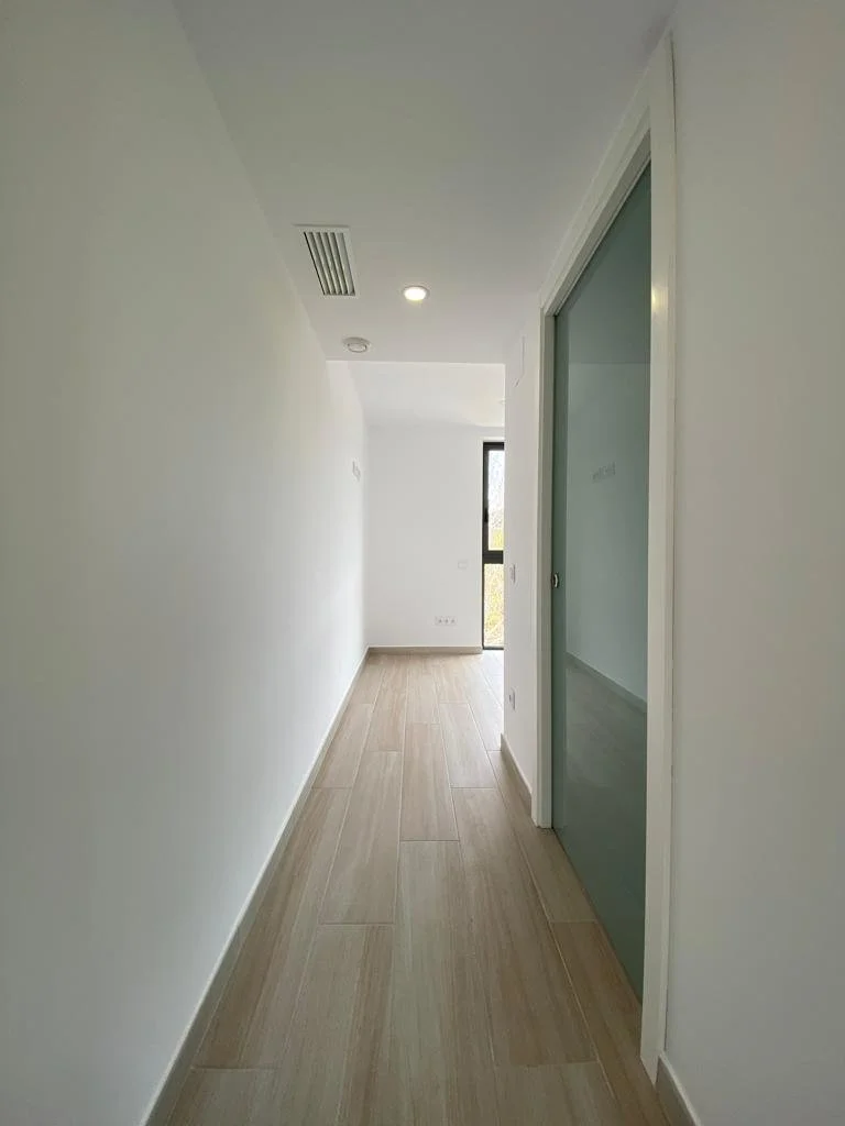 Pasillo interior en una casa moderna con paredes blancas y suelo de madera clara, con una ventana al fondo y una puerta de vidrio en el lado derecho.