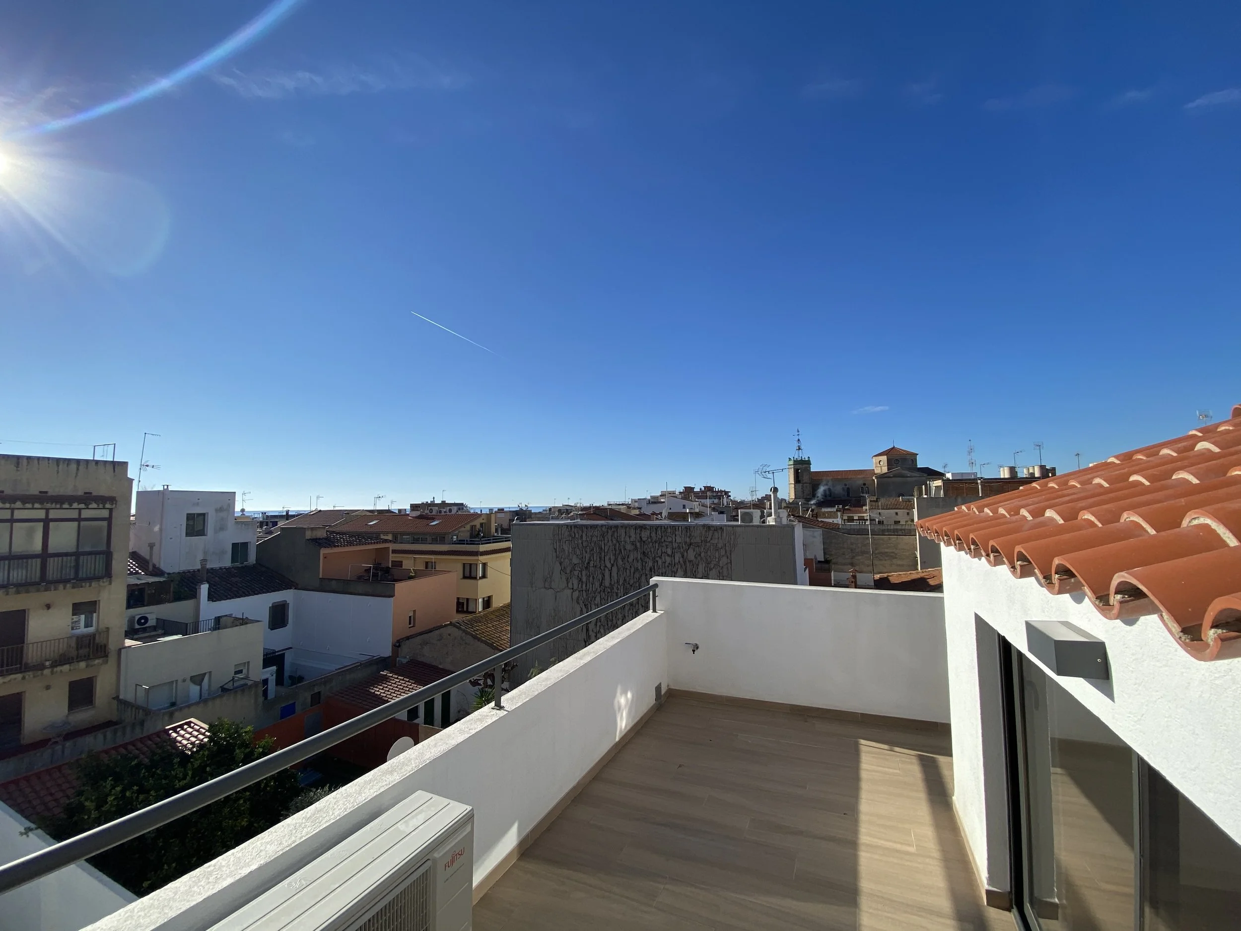 Vista desde una terraza con suelo de madera, barandilla de metal, paredes blancas, y techos de tejas rojas, observando un cielo azul con sol brillante y algunas construcciones urbanas en el fondo.