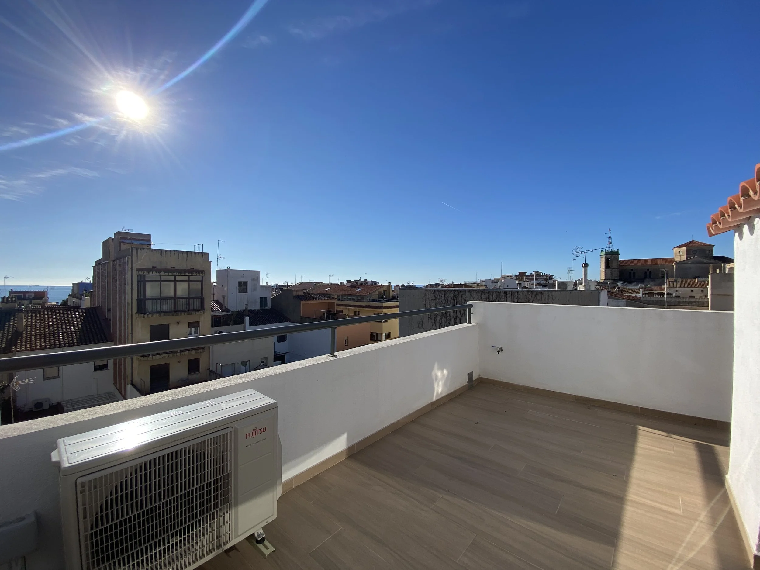 Vistas desde una terraza urbana con clima soleado y cielo despejado, edificios residenciales y un mar en la distancia.