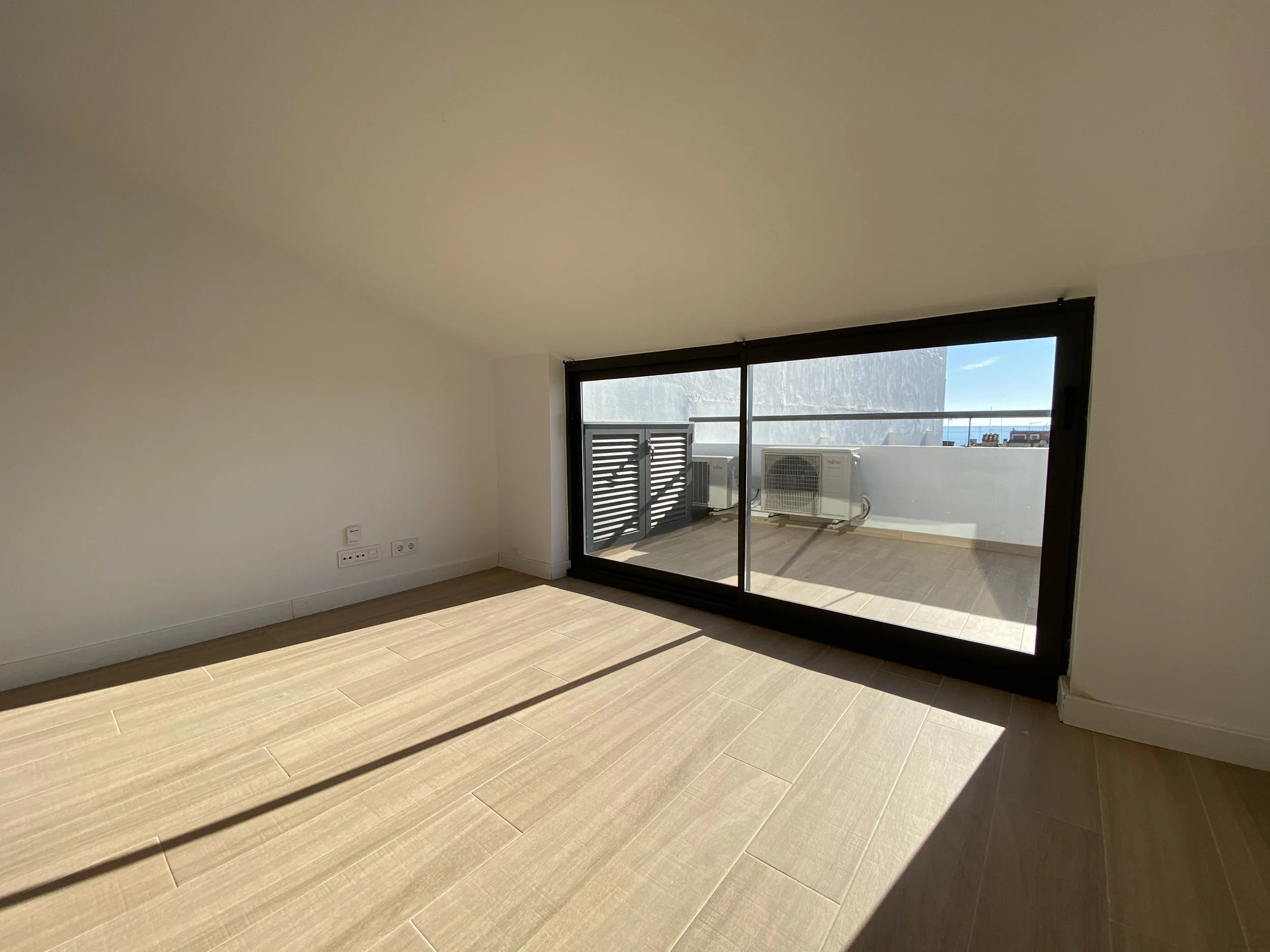Habitación vacía con piso de madera, paredes blancas y un gran ventanal que da a un balcón con aire acondicionado.