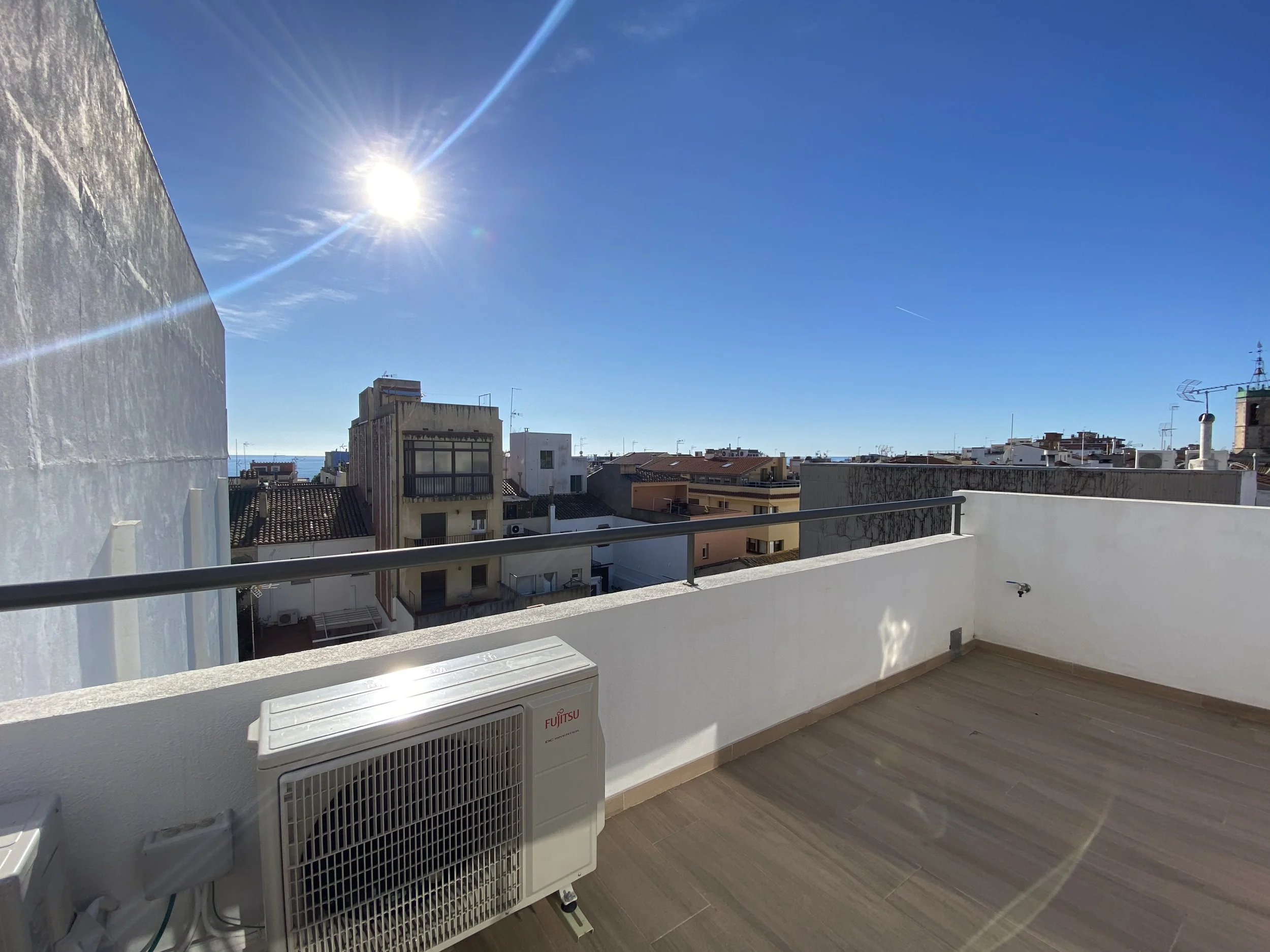 Vista desde una terraza despejada con suelo de madera, balcón con barandilla metálica, aire acondicionado en la esquina, fondo de edificios residenciales y cielo azul con sol brillante y sin nubes.