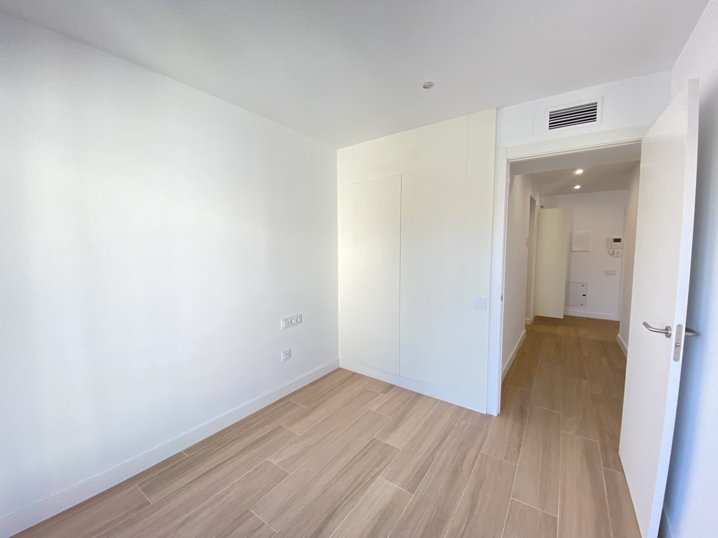 Interior de una habitación vacía con paredes blancas y piso de madera clara, con puerta abierta hacia un pasillo que también tiene paredes blancas y piso de madera.