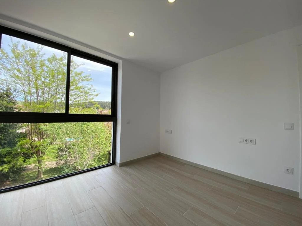 Habitación vacía con paredes blancas, suelo de baldosas claras y una ventana grande que muestra árboles y un cielo nublado.