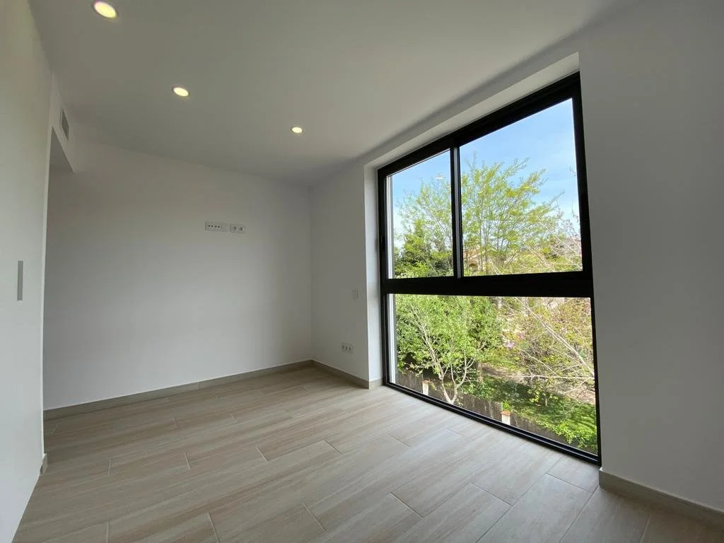 Habitación vacía con paredes blancas, suelo de madera clara, grandes ventanas de marco negro que muestran árboles verdes exteriores y cielo azul