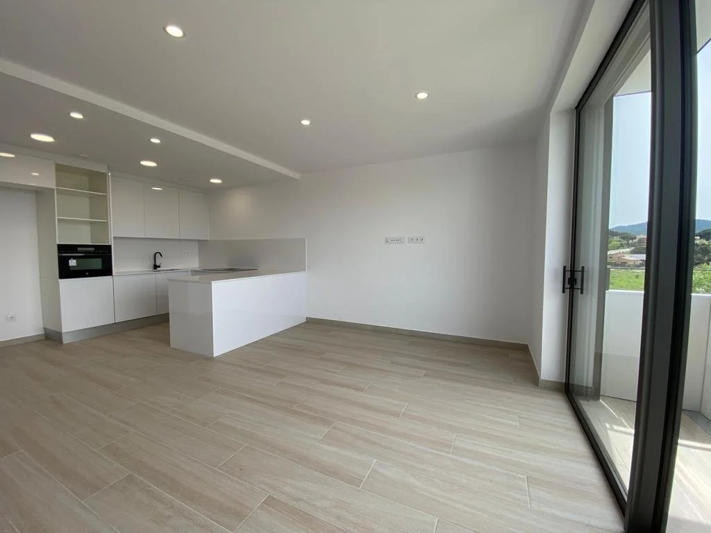 Cocina moderna con encimeras blancas, armarios blancos, paredes blancas, suelo de baldosas beige, con ventana grande de cristal que da a un balcón con vista a áreas verdes y colinas.