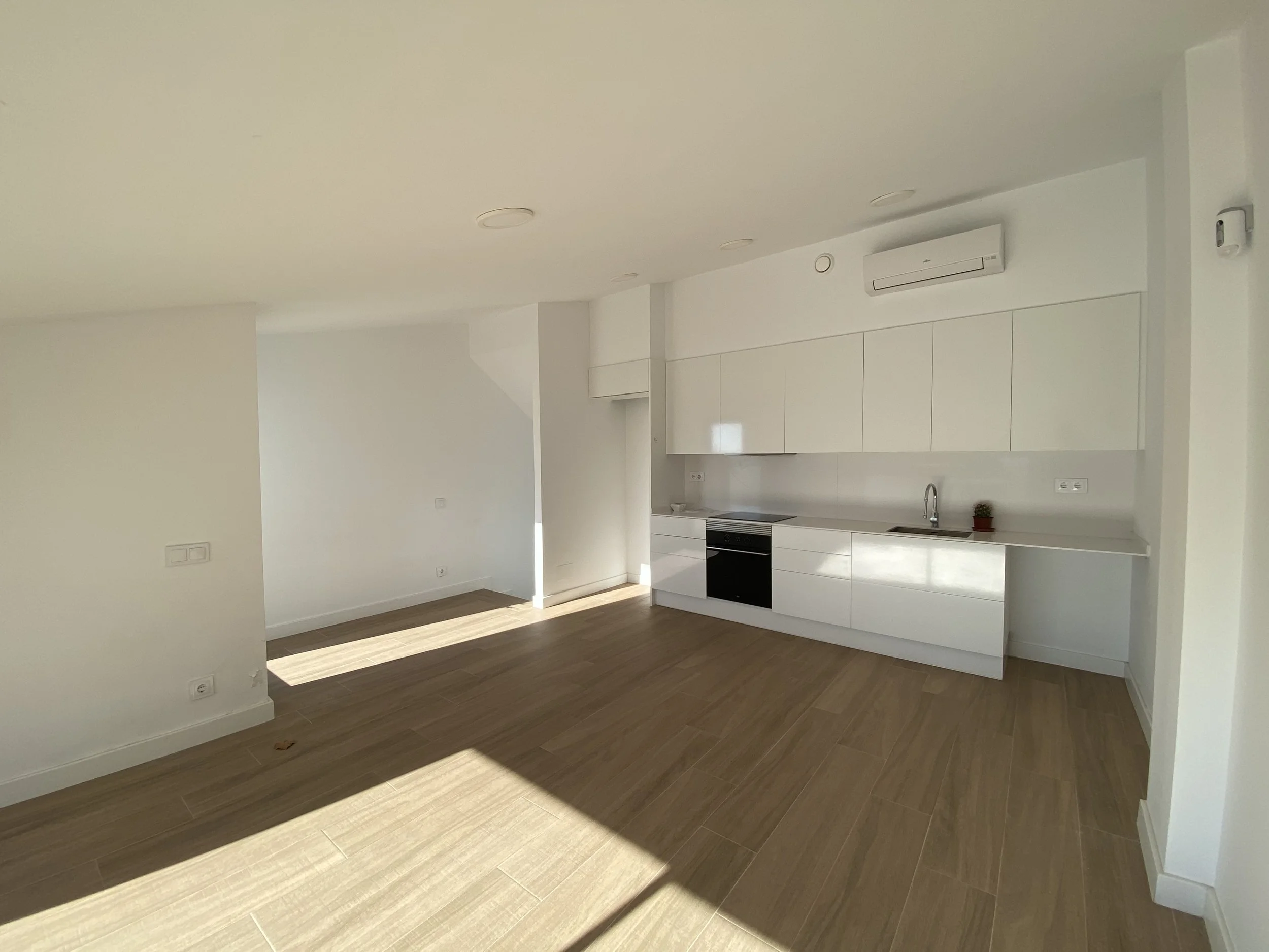 Espacio vacío de apartamento con cocina blanca moderna y suelo de madera clara, iluminado por luz natural.