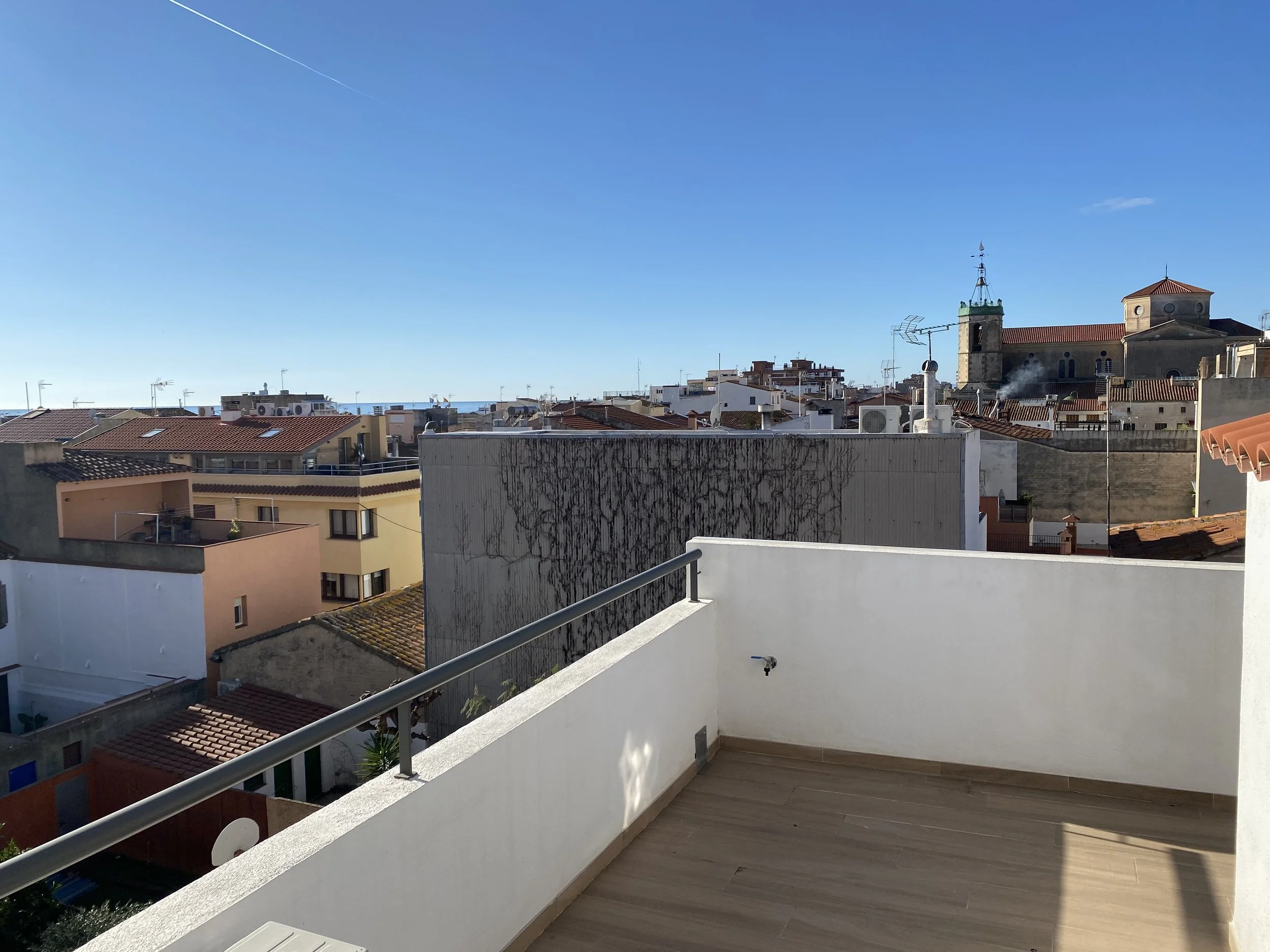 Vista desde una terraza urbana con barandilla metálica y paredes blancas, mostrando edificios con techos de tejas y antenas, una iglesia con torre y campanario, y el mar en el horizonte bajo cielo azul