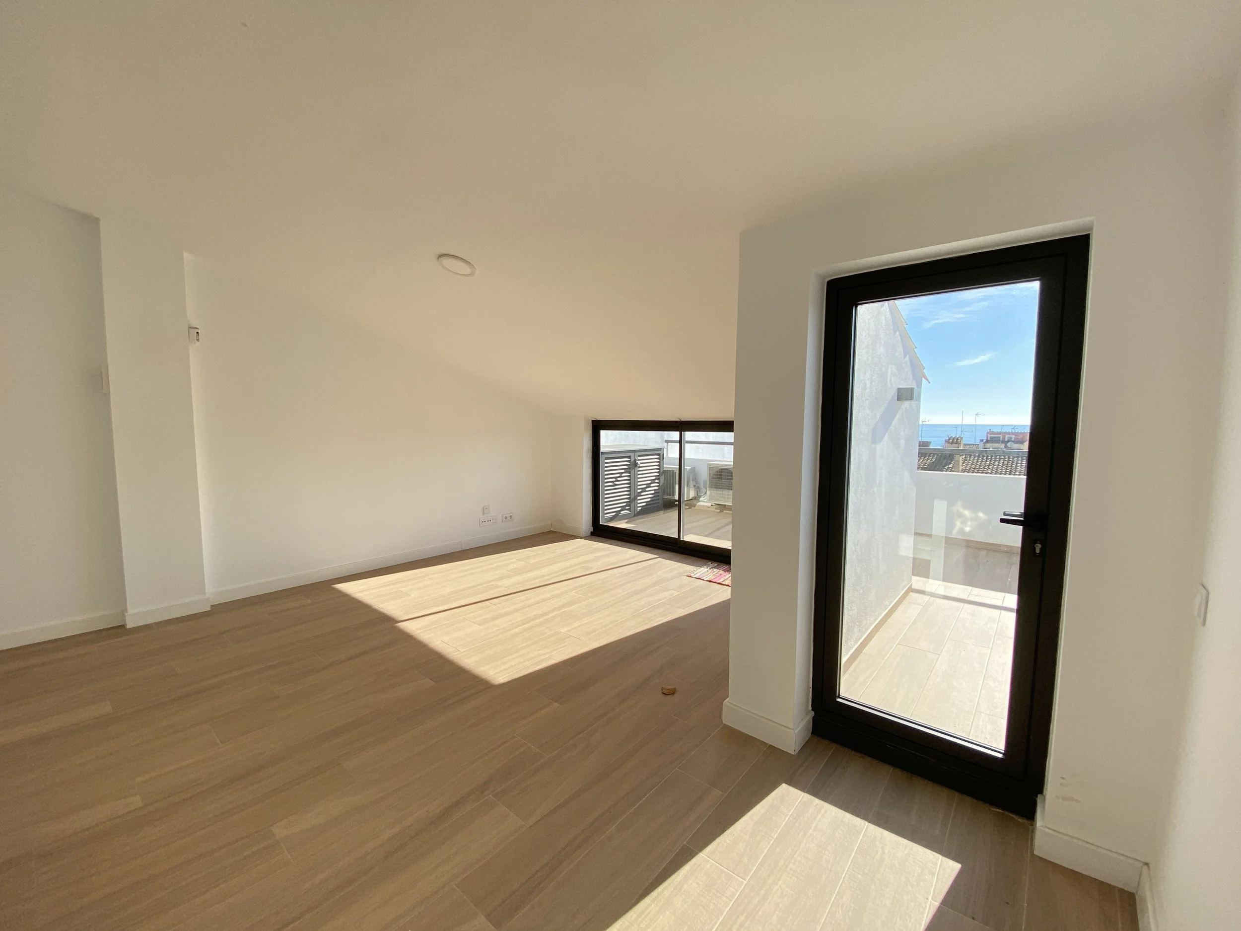 Amplio espacio interior con piso de madera y paredes blancas, con una puerta de vidrio que da a un balcón y una ventana grande que deja entrar mucha luz natural.