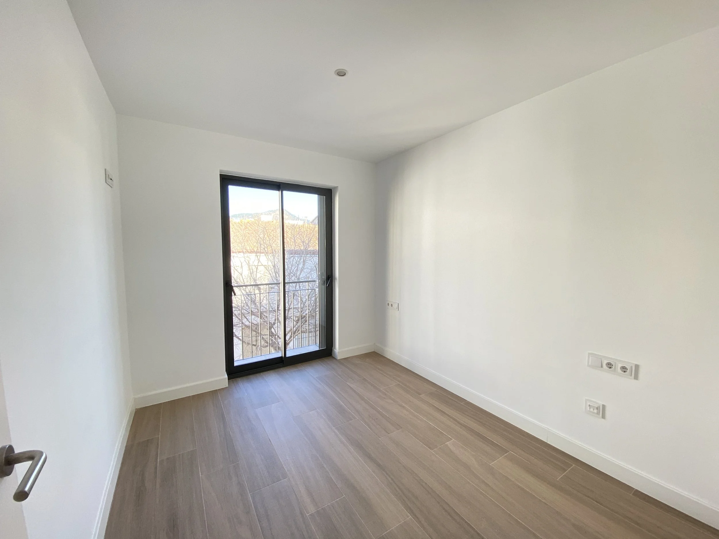 Habitación vacía con paredes blancas, suelo de madera clara, ventana de balcón con puertas correderas y barandilla, luz natural, varias tomas eléctricas en la pared.