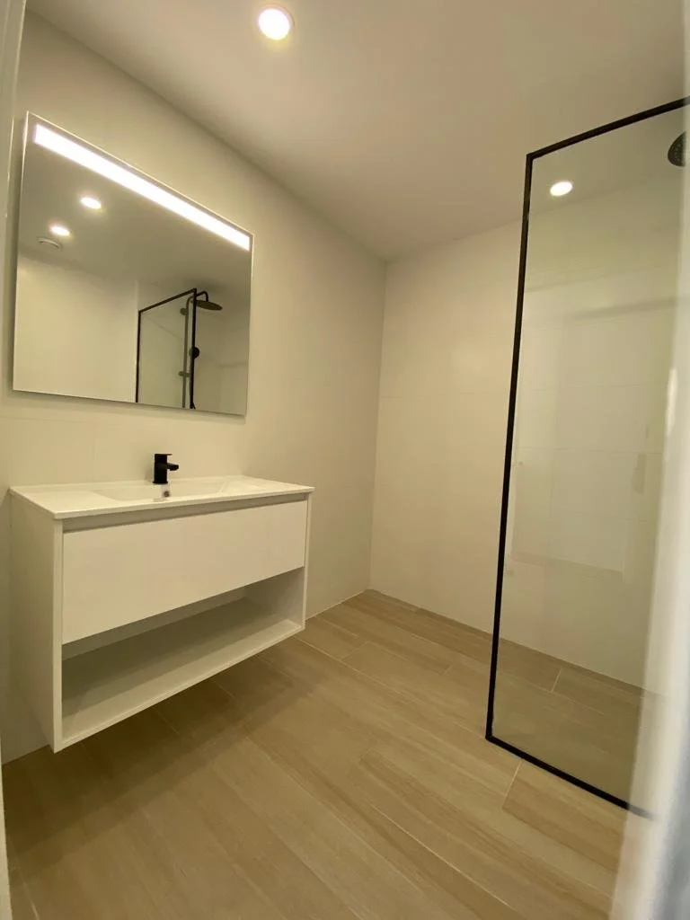 Baño moderno con lavabo blanco, espejo grande con luz LED, ducha con paredes de vidrio y piso de madera clara.