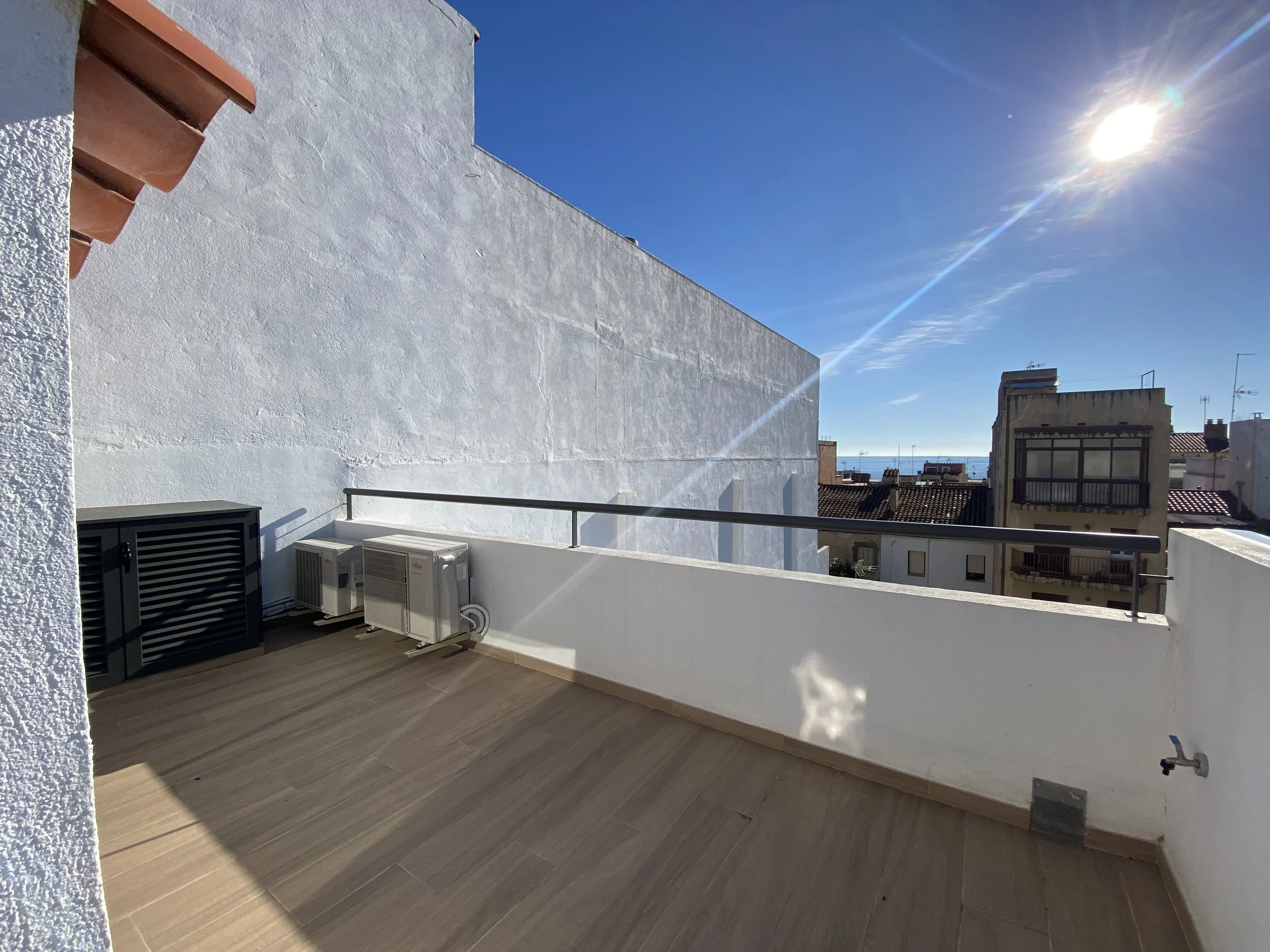Amplia terraza con suelo de madera y aire acondicionado, vistas a edificios y al mar bajo un cielo despejado y soleado.