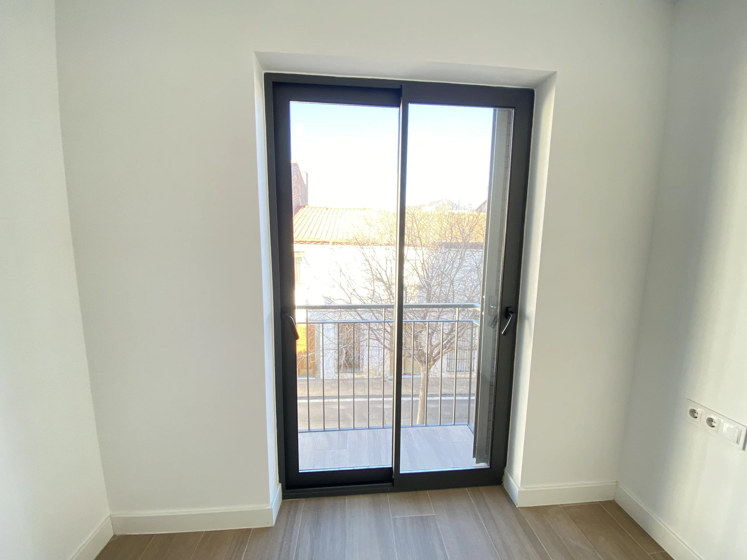 Puerta de cristal con marco negro que da a un pequeño balcón con barandilla metálica, en una habitación vacía con paredes blancas y suelo de madera.