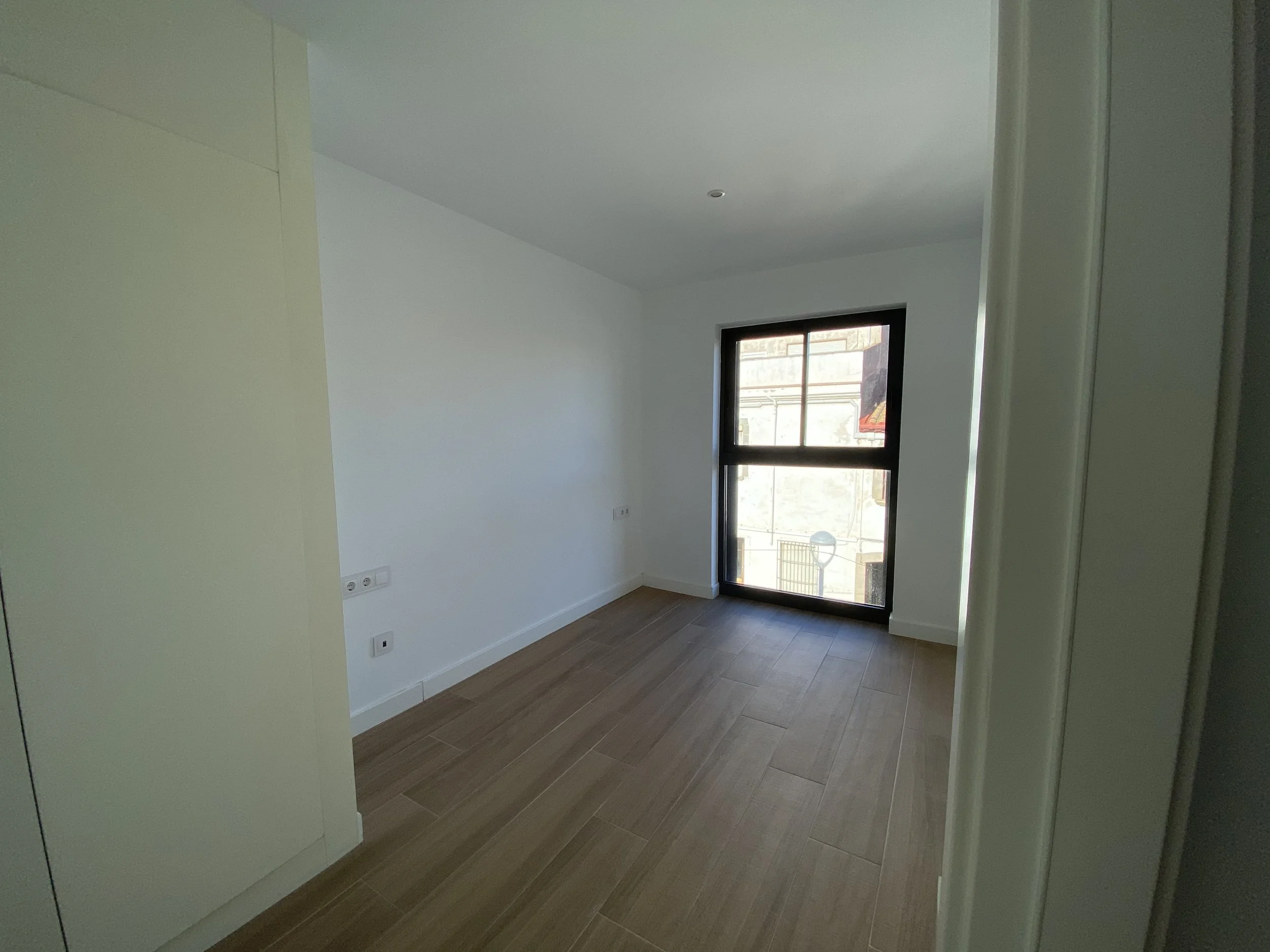 Habitación vacía con piso de madera, paredes blancas, una ventana grande con marco negro y vista a edificaciones exteriores.